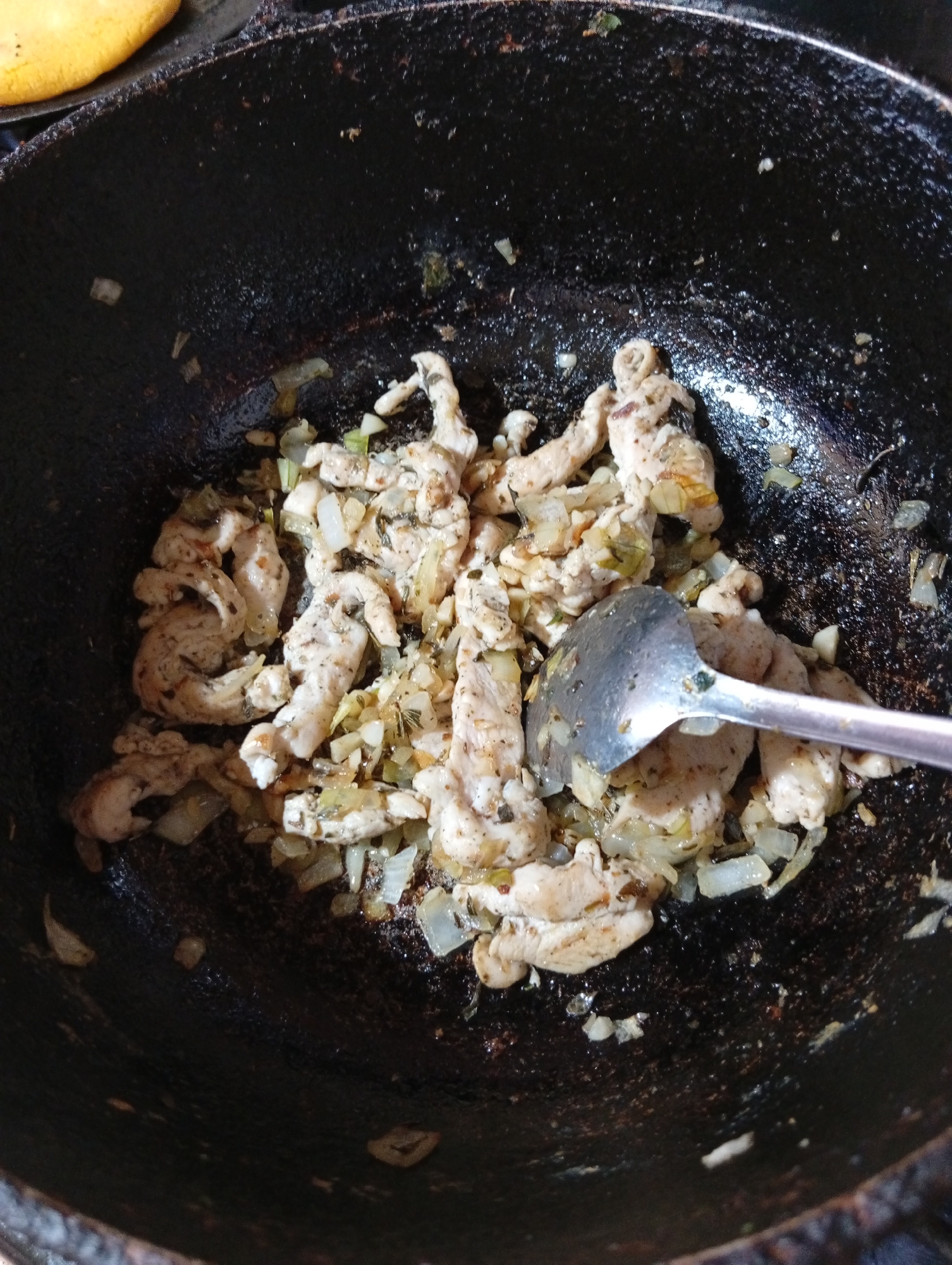 In a close-up, overhead shot, strips of cooked chicken and diced onions are being stirred in a dark, seasoned cast-iron skillet, likely in San Antonio de Los Altos, Venezuela. A silver serving spoon is partially submerged in the food, indicating an active cooking process. The skillet's surface is well-used, with a patina suggesting frequent use for preparing traditional Venezuelan dishes. The background is dark, with a blurred, yellowish object, possibly a piece of arepa or corn cake, visible in the upper left corner. The lighting is somewhat dim, casting shadows that enhance the textures of the food and the skillet.