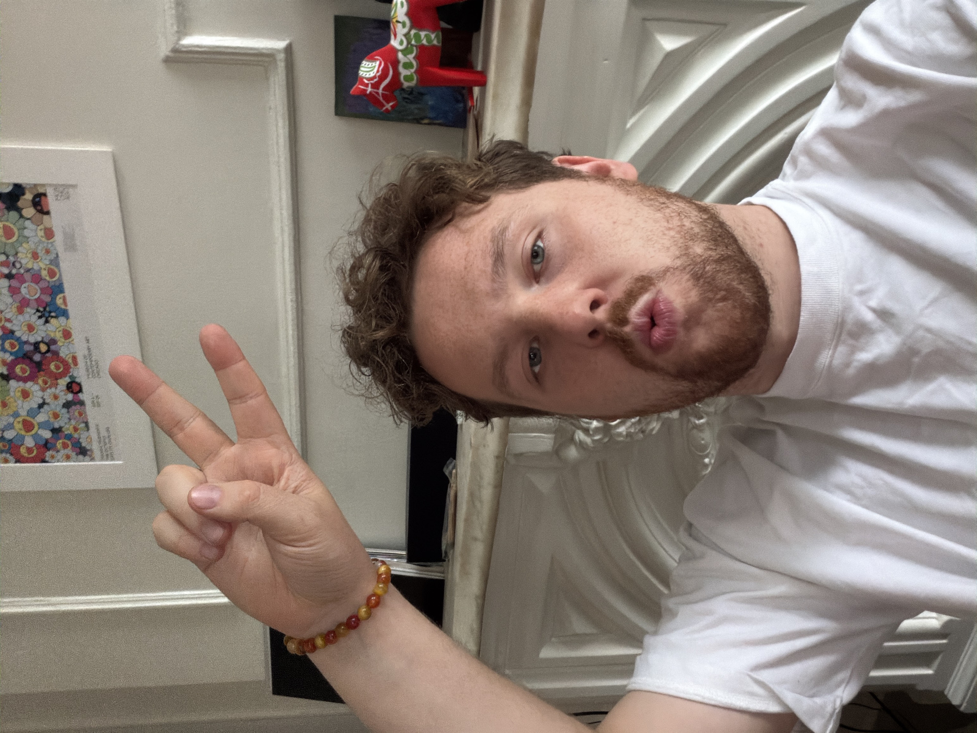 A man is posing for a selfie. He is wearing a white T-shirt and has curly brown hair and a reddish-brown beard. He is making a "peace" sign with his left hand, and his face is tilted towards the camera with pursed lips. He is wearing a beaded bracelet on his left wrist.
Behind him are a framed piece of art with colorful floral designs, a small red horse figurine, and a white door with decorative molding. The location is likely inside a residential building. There is no information available in the image about the specific location in the city of New York.