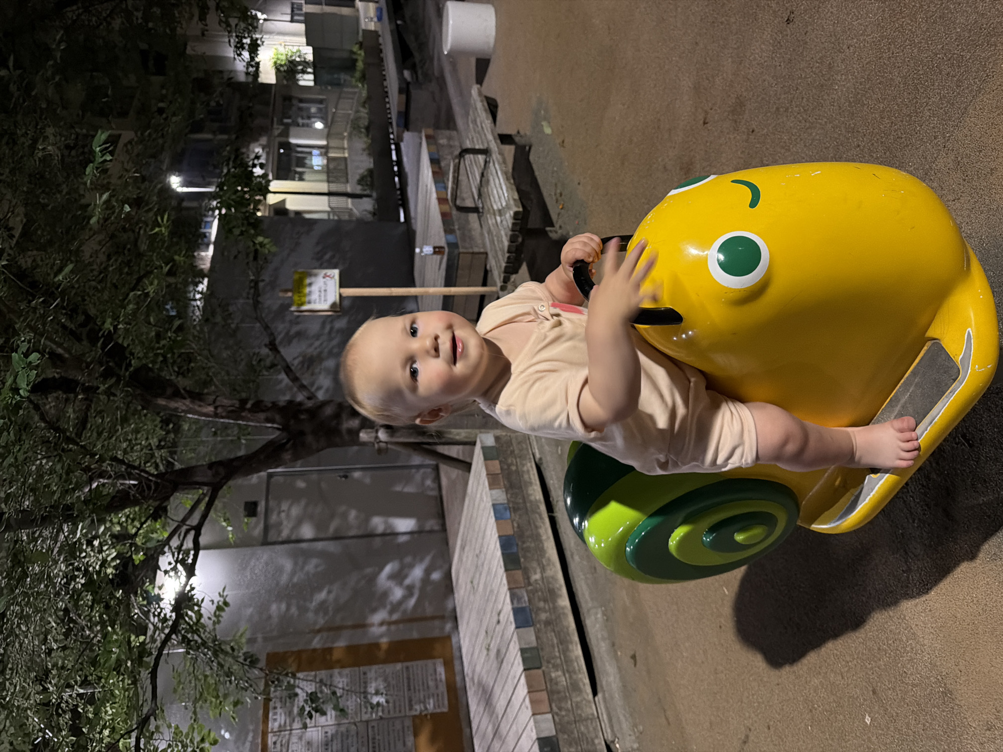 In the city of Chuo, Japan, at night, a young child sits on a yellow snail-shaped play ride in a park. The child is wearing a light peach-colored onesie and is facing the camera with a slight smile. The snail ride has green details on its shell and large green eyes. The background shows trees with visible leaves, a building, and park benches. The ground appears to be covered in small pebbles or gravel.