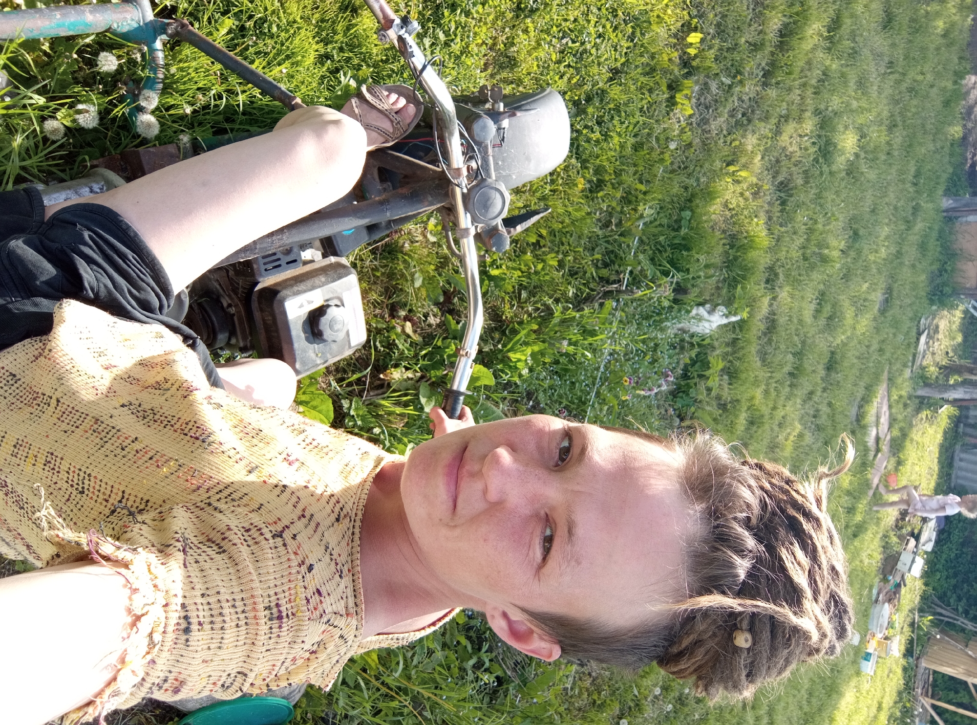 The image captures a person taking a selfie outdoors in a sunny, grassy environment, likely a garden or backyard. The camera is held by the subject, providing a slightly downward view of their upper body and surrounding elements.
The central subject is a person with fair skin, freckles, and brown eyes, displaying a neutral to gently smiling expression. Their hair is styled in dreadlocks, with some pulled back into a bun and the sides appearing very short or shaved. They are wearing a light-colored, possibly yellow or beige, knitted or crocheted top adorned with multicolored flecks. Their legs are visible, and they have sandals on their feet.
The person is seated on or next to a small motorized vehicle, resembling a mini-bike or scooter. Visible parts include a small engine with a black fuel cap situated between their legs, chrome handlebars equipped with black controls, and a segment of a turquoise or light blue metal frame near their left arm.
The setting is an open-air area lush with green grass, dotted with several white dandelion seed heads. The bright sunshine suggests a clear day, likely late morning or afternoon, as evidenced by strong highlights and shadows. In the distant background, a blurry figure, possibly a child, is visible further into the grass, along with glimpses of a wooden fence or structure and various miscellaneous garden items.
The image depicts a casual outdoor moment. The location is Ostrovtsy, Russia. No visible text is present.