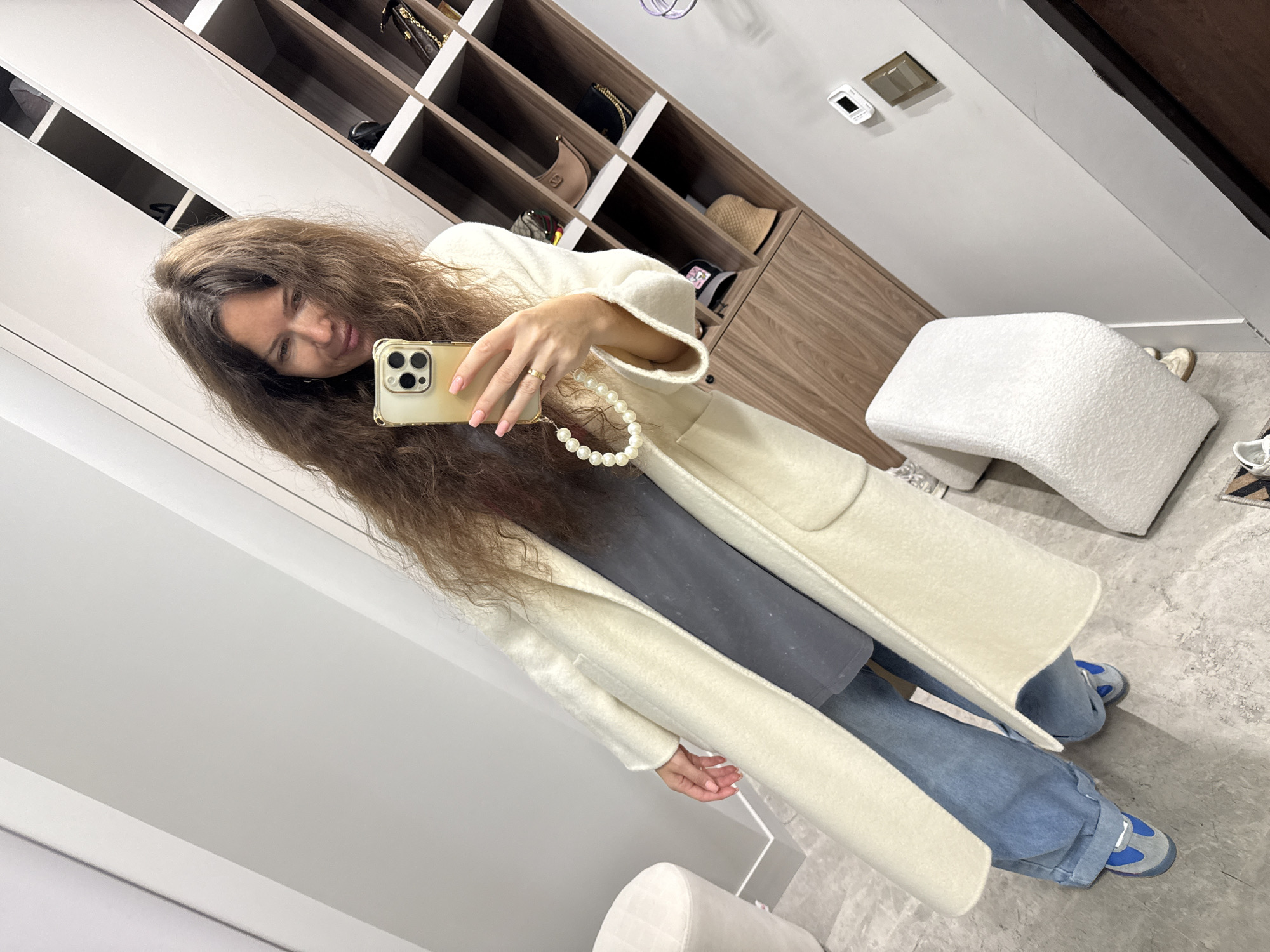 A woman with long, wavy brown hair is taking a mirror selfie in what appears to be a walk-in closet or dressing room. She is wearing a long, cream-colored coat over a dark gray t-shirt and light blue wide-leg jeans. On her feet are gray and blue sneakers. Her right hand is holding a gold-colored smartphone, which has a pearl-beaded strap attached. She is wearing a thin gold ring on her left ring finger. The background shows shelves filled with handbags and other accessories. A white textured ottoman is visible to the left of the woman, and a white, curved armchair is in the foreground on the right. The overall environment suggests a modern and organized personal dressing space.