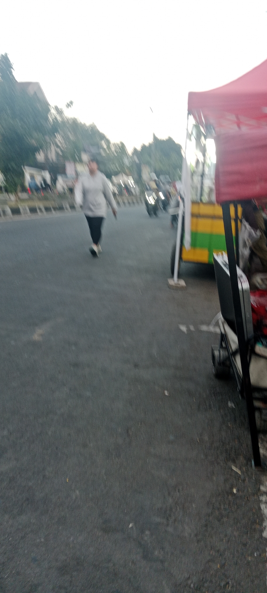 A slightly blurry, medium shot depicts an outdoor scene during the day on a street in Ngemplak, Indonesia. The focus is on a person in the mid-ground walking on the asphalt road. The individual is wearing a light-colored, possibly gray, long-sleeved shirt and dark pants. They are facing the camera and appear to be in motion. To the right of the road, a portion of a red tent and a yellow and green striped cart or structure are visible. In the background, there are buildings, trees, and a few vehicles, including what appears to be a motorcycle. The overall impression is of a street scene with pedestrian activity and possibly a vendor's stall.