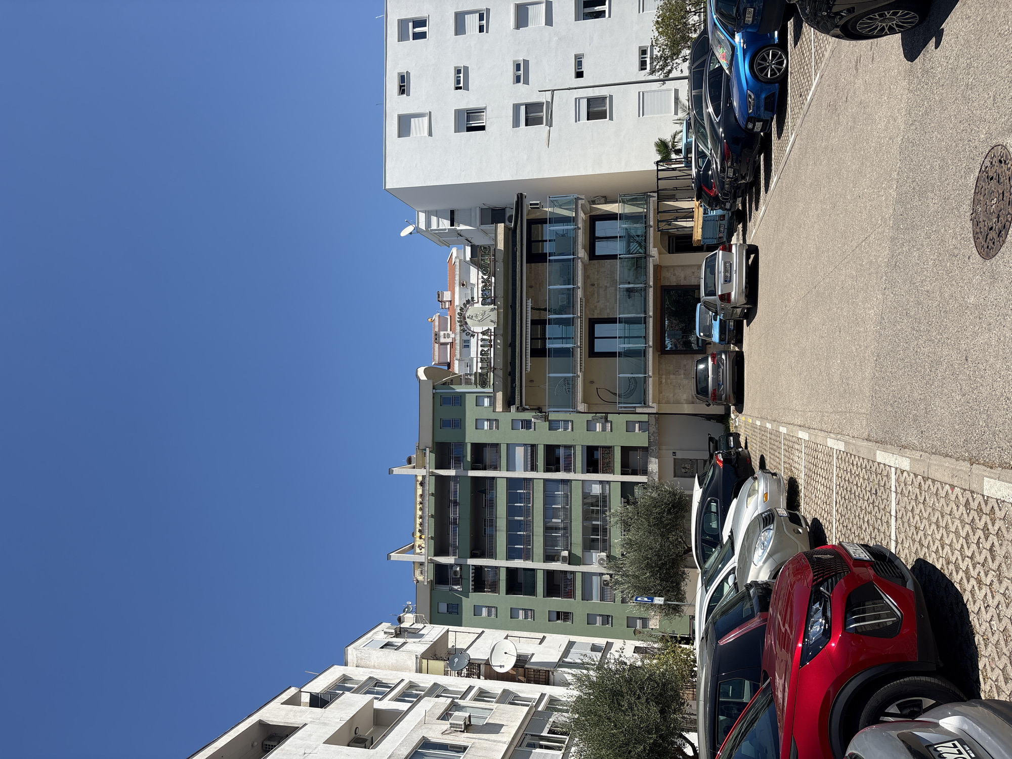 This is an exterior, eye-level, vertical, medium shot of a parking lot in front of several buildings under a clear blue sky. In the foreground, the curb and a textured, grey, paved parking area are visible, with several cars parked in designated spots. To the left, a red Peugeot 3008 SUV is parked in the closest spot, facing right. Further back in the parking lot, a row of cars includes a black SUV, a silver sedan, and a black sedan with a spoiler. On the right side of the parking lot, a blue Audi A3 sedan is parked, followed by other dark-colored vehicles.
The background features a row of multi-story buildings. The closest building on the right is a white, modern structure with numerous windows, some with closed white shutters. Adjacent to it is a building with a beige lower section and a darker upper section featuring balconies with glass railings. Above this, a green, multi-story building with balconies and many windows dominates the center of the frame. Further left, a white building with a more complex facade and visible air conditioning units is also present. Trees with sparse, green foliage are visible in front of the buildings. A blue parking sign with a white "P" is mounted on a pole between the green and white buildings. The buildings appear to be residential or commercial, with balconies and satellite dishes visible. The overall scene depicts an urban environment on a sunny day.