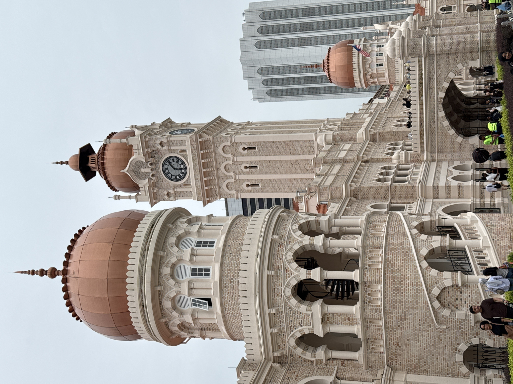 The image shows the Sultan Abdul Samad Building in Kuala Lumpur, Malaysia. It is a late nineteenth century building located along Jalan Raja in front of the Merdeka Square. The building is made of red brick and features Moorish architecture, including arches, domes, and a clock tower. The clock tower has a clock face displaying the time. There are people visible around the building. A modern high-rise building is also visible in the background. The sky is overcast.