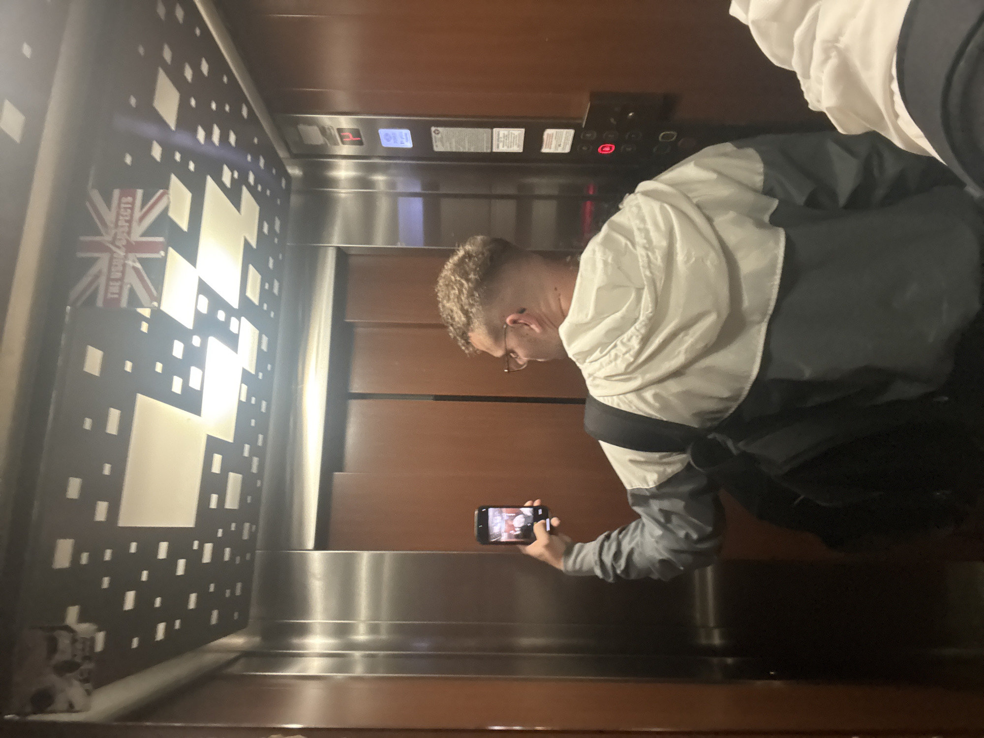 The image depicts the interior of an elevator in Plovdiv, Bulgaria, with a person in the foreground capturing a moment with their smartphone. The photograph is oriented vertically, with the elevator floor at the bottom and ceiling at the top, taken from a slight angle.
The main subject is a male individual, seen from the side, wearing glasses and a multi-colored jacket (white, grey, and black sections). He has short, curly, light-colored hair. He holds a black smartphone horizontally, seemingly taking a picture of his reflection or the elevator's interior directly in front of him. A partial view of another person, showing the top of a head and a white hood, is visible on the far right edge of the frame.
The elevator's interior walls are distinct. One side features warm wooden paneling. The opposing wall, which is more prominent, is dark with a decorative pattern of numerous small and large light-colored rectangular and square shapes. A central design element on this wall is a stylized Union Jack flag, below which the text "THE USUAL SUSPECTS" is clearly visible in red. The lower part of the elevator walls and the base feature reflective stainless steel.
An elevator control panel is visible on the wooden wall, displaying "H" for the current floor and a red illuminated "1" likely indicating the destination floor. The lighting inside the elevator appears bright and artificial. The scene captures an ordinary moment of someone taking a photo within a uniquely decorated elevator space.
