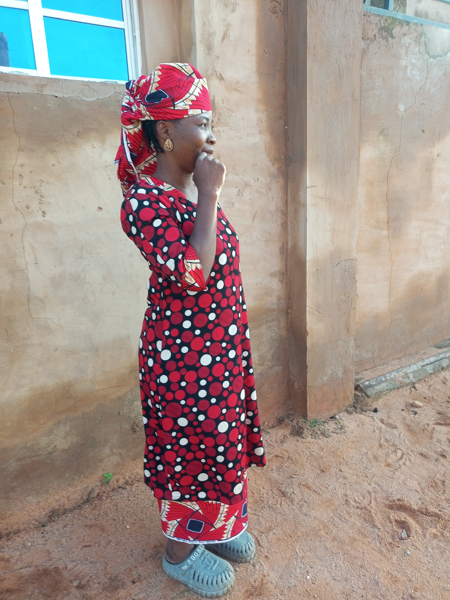 A medium shot captures a woman standing outside a building in Tempure, Nigeria. She is wearing a red and black patterned dress with white polka dots and a matching headscarf. The woman is looking to her right with her hand on her chin. The building behind her is constructed of rough, light brown concrete with some cracks and stains. A window with blue panes is visible to her left. The ground is covered in reddish-brown sand.