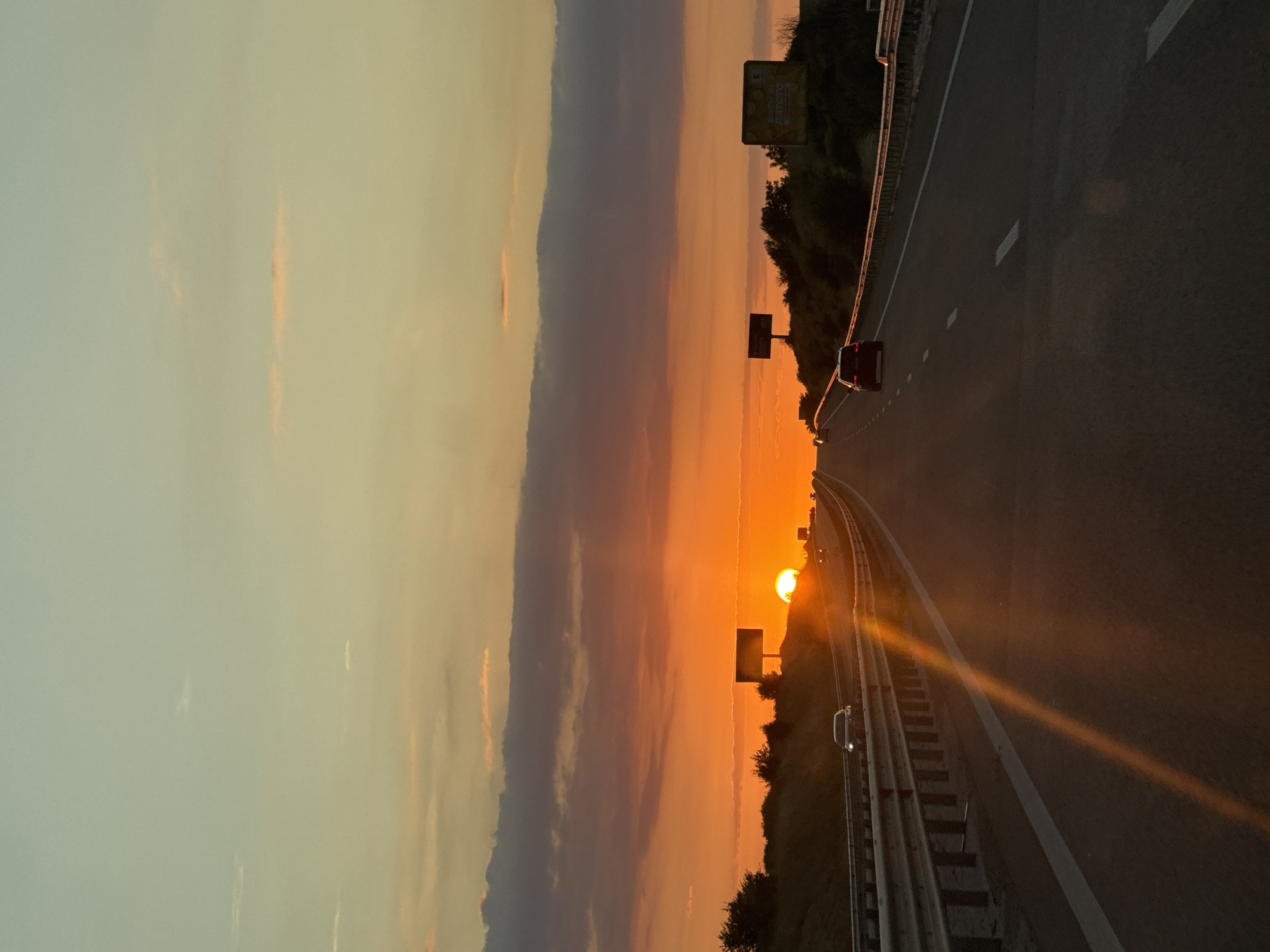 The image depicts a highway in the city of Yazd, Bulgaria, at sunset. The sky is filled with vibrant orange and yellow hues, with some darker clouds present. The sun is low on the horizon, casting a warm glow and creating lens flare effects on the road. Several cars are visible on the highway, traveling in both directions. Guardrails line the sides of the road, and there are billboards visible in the distance. The landscape appears to be semi-arid, with some sparse vegetation on the hillsides. The overall impression is one of a peaceful and scenic drive during twilight.