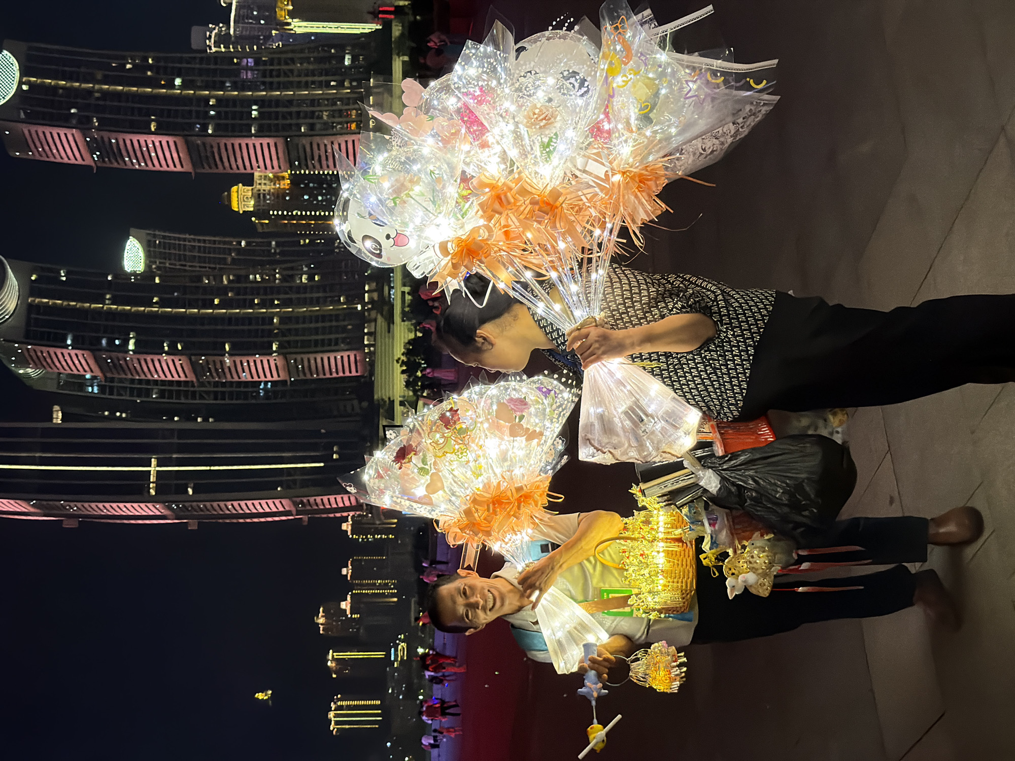 In Jiangbeicheng Street, China, at night, a man stands holding a basket of illuminated decorations and a toy airplane. He smiles at the camera, his basket filled with golden lights. To his right, a woman holds a large bouquet of illuminated balloons, some adorned with cartoon faces like pandas. The background features towering, modern buildings lit with pink and yellow lights, creating a vibrant urban cityscape. People can be seen in the distance, walking and enjoying the evening. The scene depicts a lively street market atmosphere with vendors selling light-up novelty items.