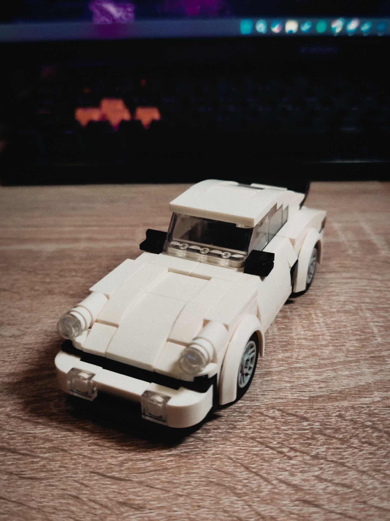 A toy car made of white Legos sits on a wood-grain table. The toy car has a distinctive sports car shape, with rounded fenders and a spoiler. In the blurred background is a laptop. The location is not identified.