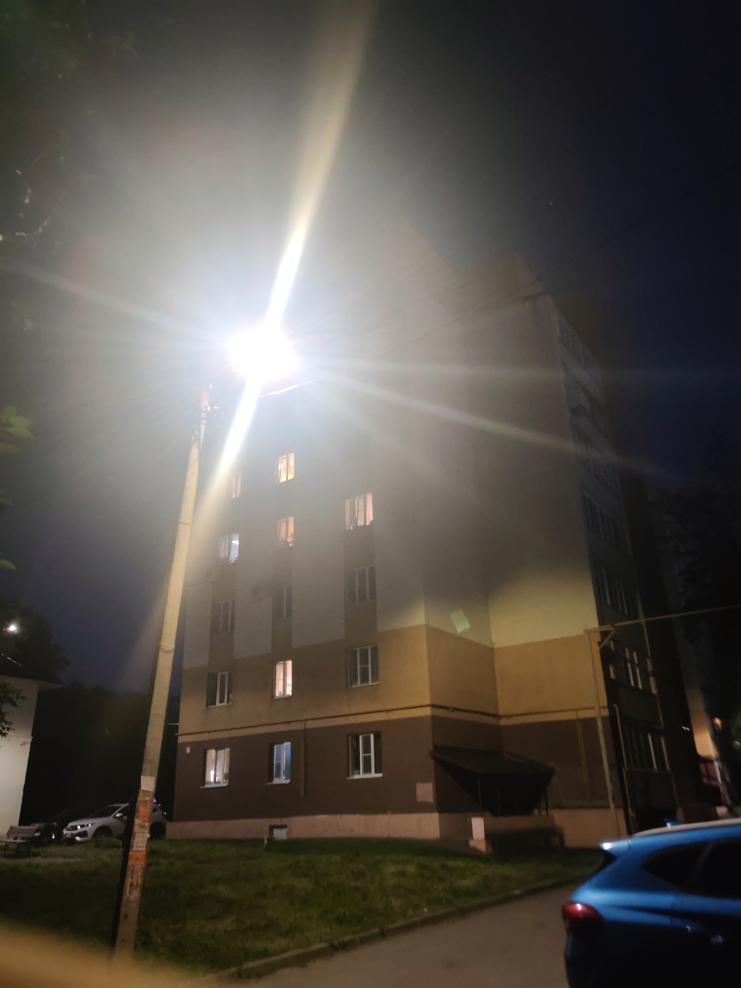 The image captures a nighttime scene in a residential area. A tall, multi-story building with illuminated windows dominates the frame. A bright streetlight with a visible lens flare is positioned to the left of the building, casting a strong light that washes out parts of the scene and creates lens flares. In the foreground, a blue car is partially visible on the right side of the image. To the left, a patch of grass and a sidewalk are visible, with a parked white car and a bench also present. The sky is dark, indicating it is nighttime. The architectural style and the presence of a streetlight suggest an urban environment. Based on visual cues, this scene is consistent with a typical residential complex in a city like Vladimir, Russia.