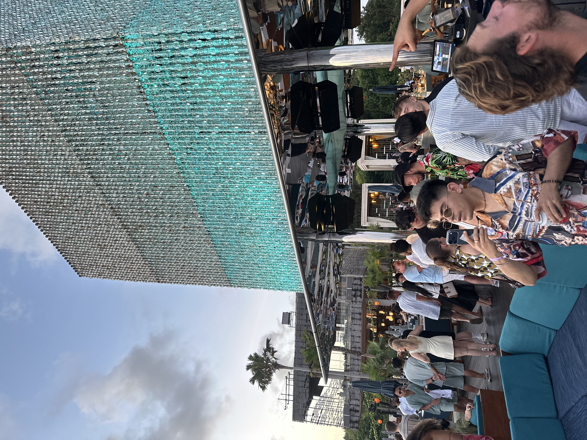 The image depicts a gathering of people on a rooftop bar or similar venue in Kuta Selatan, Indonesia. The location appears to be elevated, as evidenced by the view of the sky, clouds, and a palm tree in the background. The setting suggests a social event or party with people casually dressed and interacting with each other. Some individuals are taking pictures with their phones. There are blue cushions on the chairs and couches of the establishment. Overhead, a structural feature, possibly a decorative element, is constructed from a grid of silver and turquoise beads or similar material. It reflects the people and the sky. There are also elements of the bar's structure reflected in the metal support above the gathering of people.