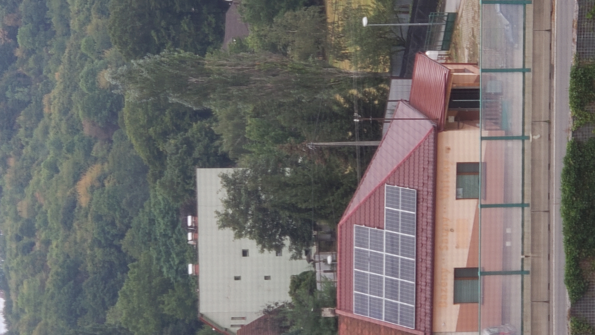 The image shows a view of a residential building with a red tiled roof covered in solar panels, located in the city of Prešov, Slovakia. The building has a beige and light brown facade with two green-shuttered windows and text that appears to read "Bazény...Sauny...Ubytko". A clear glass and green metal fence runs in front of the building, obscuring a road below. In the background, a larger, light-colored building with a flat roof and small square windows is visible, set against a backdrop of densely forested hills. The sky is overcast.