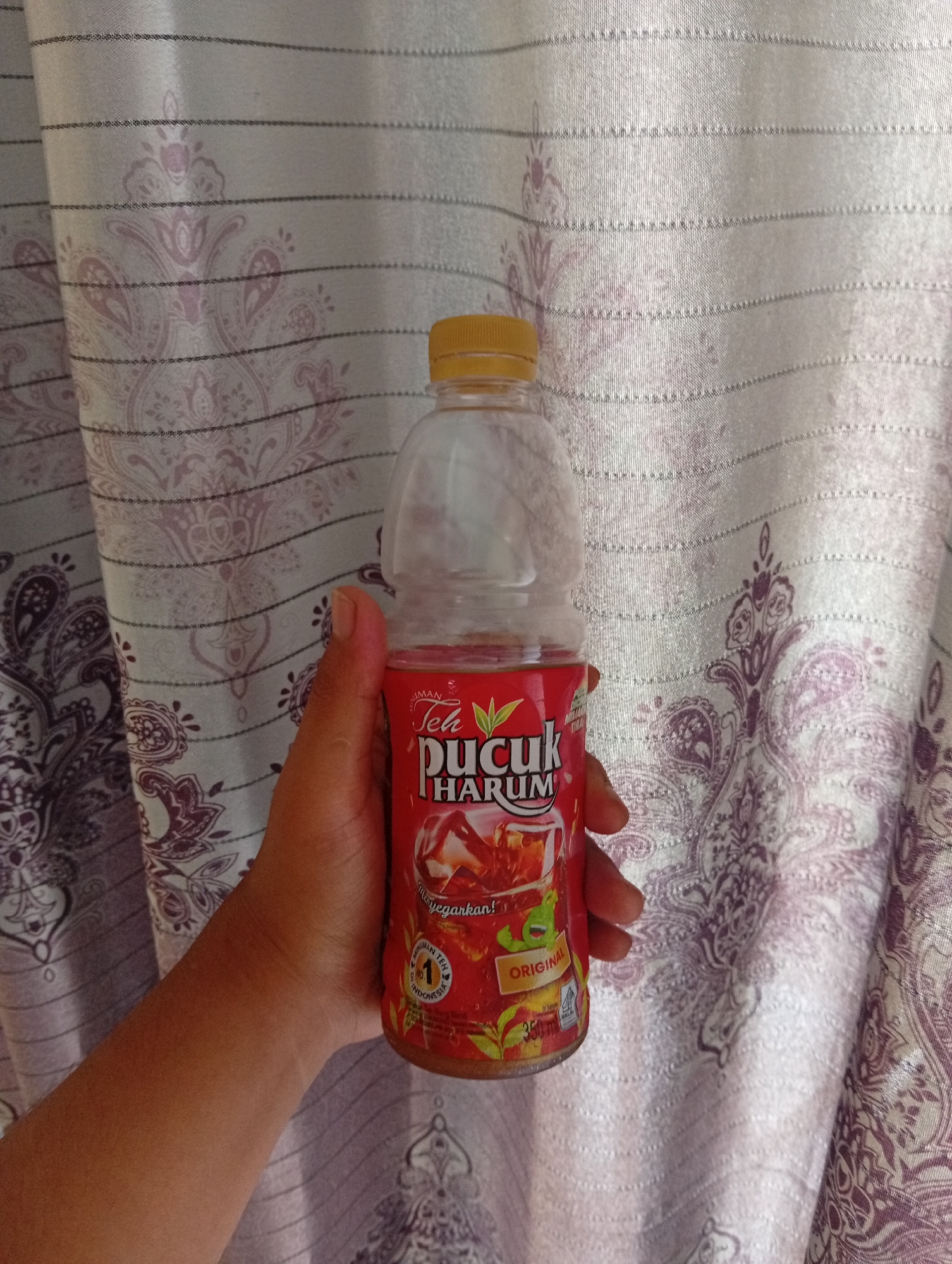A hand holds a plastic bottle of Teh Pucuk Harum, an original flavored iced tea, in front of a light purple patterned curtain. The bottle is transparent with a red label. The hand is tanned and positioned to the left of the bottle. The background consists of a fabric curtain with a repeating paisley-like pattern in shades of light purple and white. The lighting appears to be natural, coming from the front and casting soft shadows. The scene is indoor. There is no information about the location in Kualu, Indonesia.