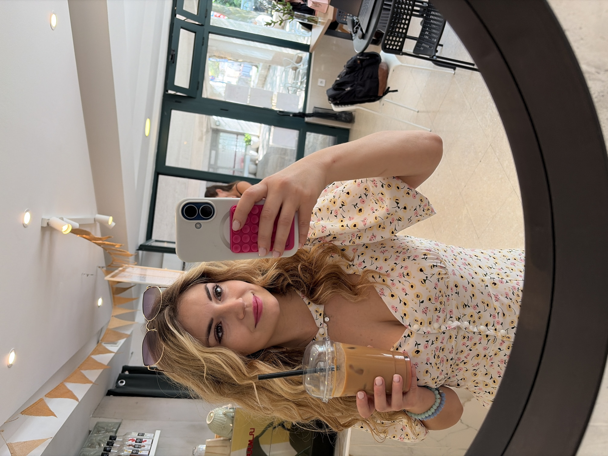 A woman with long blonde hair is taking a selfie in a mirror. She is holding a clear plastic cup with a brown beverage and a black straw. She is wearing sunglasses on her head and a floral-patterned dress with white buttons down the front. The woman is also wearing a bracelet.
The background shows a cafe interior, with a white wall decorated with a string of pennant banners. There are shelves displaying bottles and containers. A glass door and window are visible, revealing a cityscape with buildings and greenery. Another woman can be seen in the background. The location appears to be in a cafe in Budva, Montenegro. A black chair with a metal frame and a black backpack are seen in the background as well.