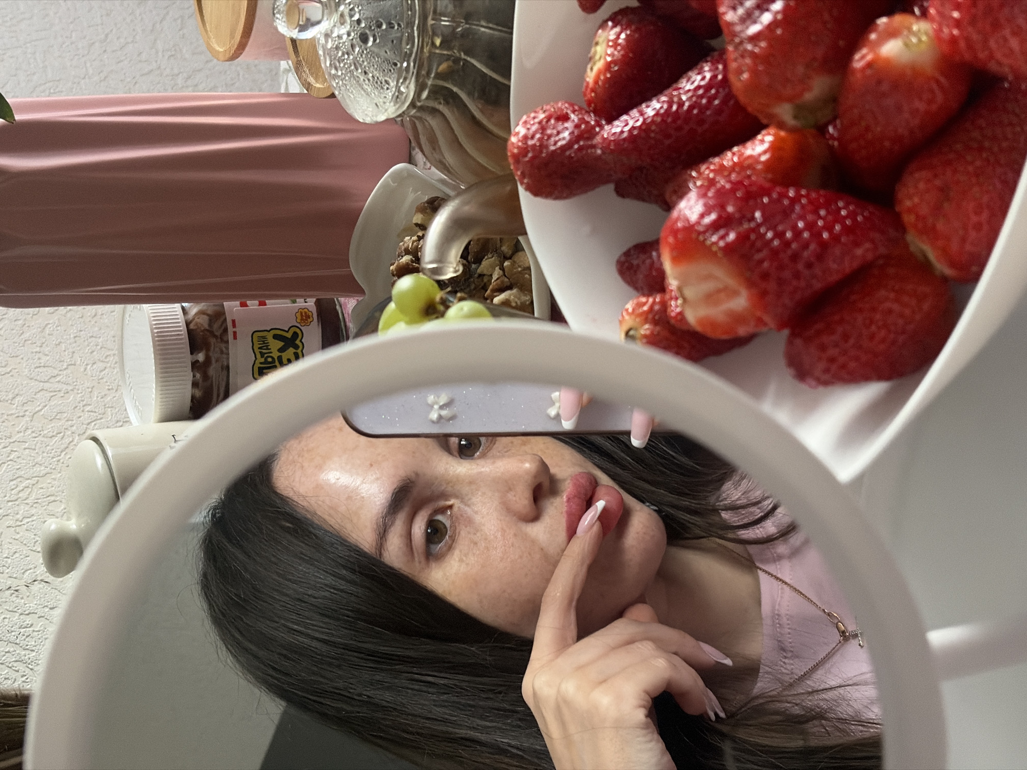 A woman is reflected in a circular mirror or ring light. She has dark hair, fair skin with freckles, and is wearing a pink top and a gold chain necklace with a small cross pendant. Her nails are painted with a French manicure. She is holding her finger to her lips. Above her reflection, a bowl of strawberries is visible, along with a jar of what appears to be a nut butter spread, a pink-wrapped object, a glass container, and a dish with walnuts and grapes. The reflection and items are located indoors in the city of Saint Petersburg, Russia.