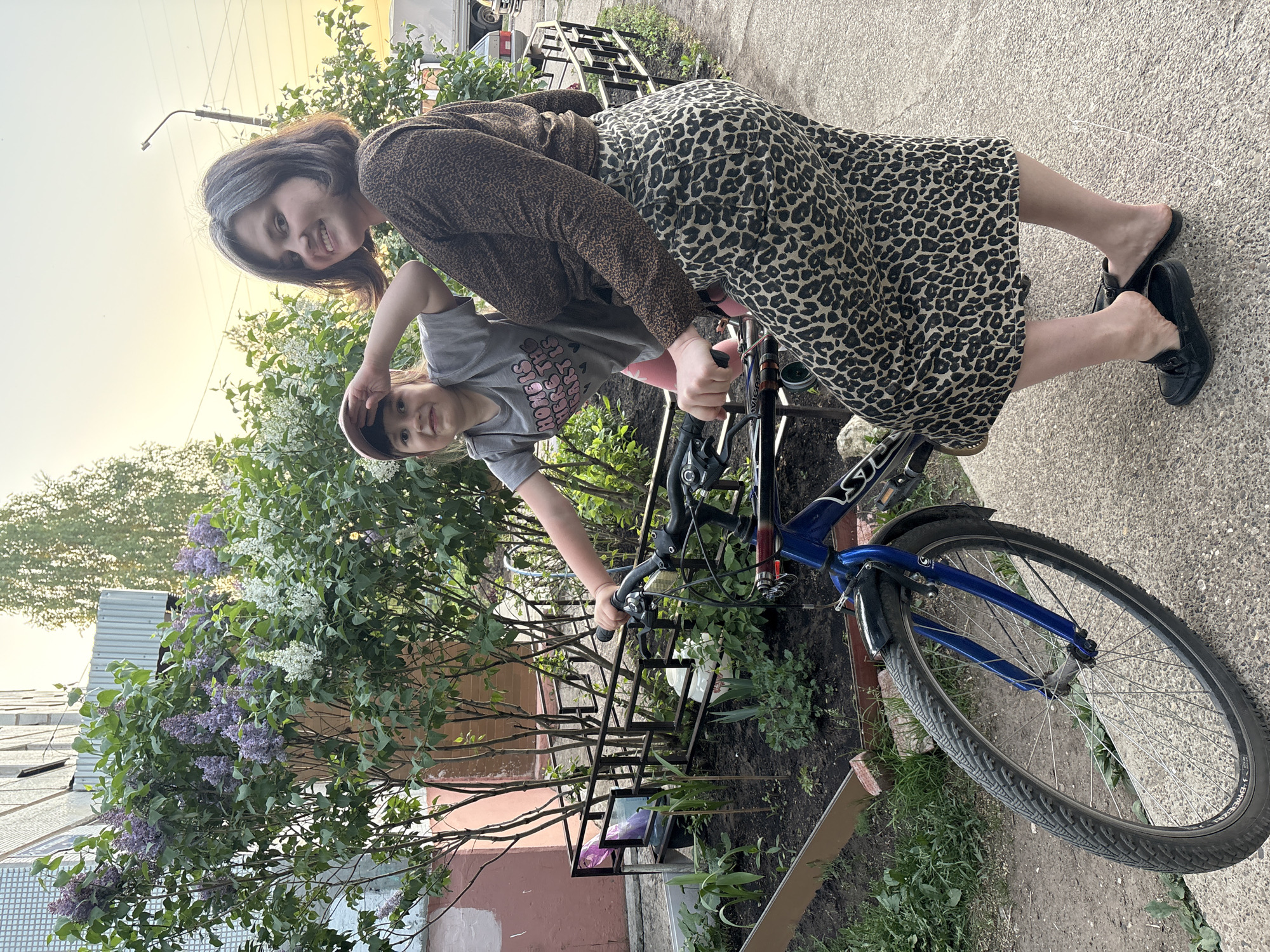 Two people are posing with a blue bicycle, one of whom is seated on the bicycle. The standing person is wearing a leopard print dress and brown shoes. The seated person is wearing a grey shirt and a hat. They are outside near a lilac bush and a building. The photograph was taken in the city of Chaikovsky, Russia.