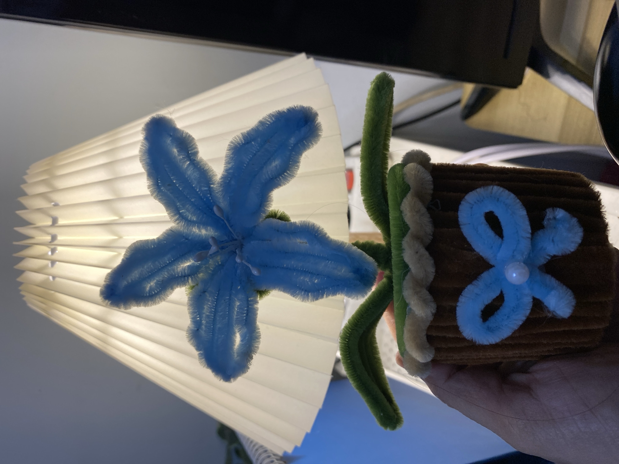 An overhead shot shows a handmade blue pipe cleaner flower in a brown pipe cleaner pot. The flower has five petals and a white center. The pot has a light brown rim and a light blue pipe cleaner bow with a white pearl in the middle. Two green pipe cleaner leaves extend from the base of the flower. The flower is held in a person's hand. Behind the flower is a white pleated lampshade. The background includes a glimpse of a desk and computer monitor. The image is lit by the lamp. This image was taken in Babelan, Indonesia.