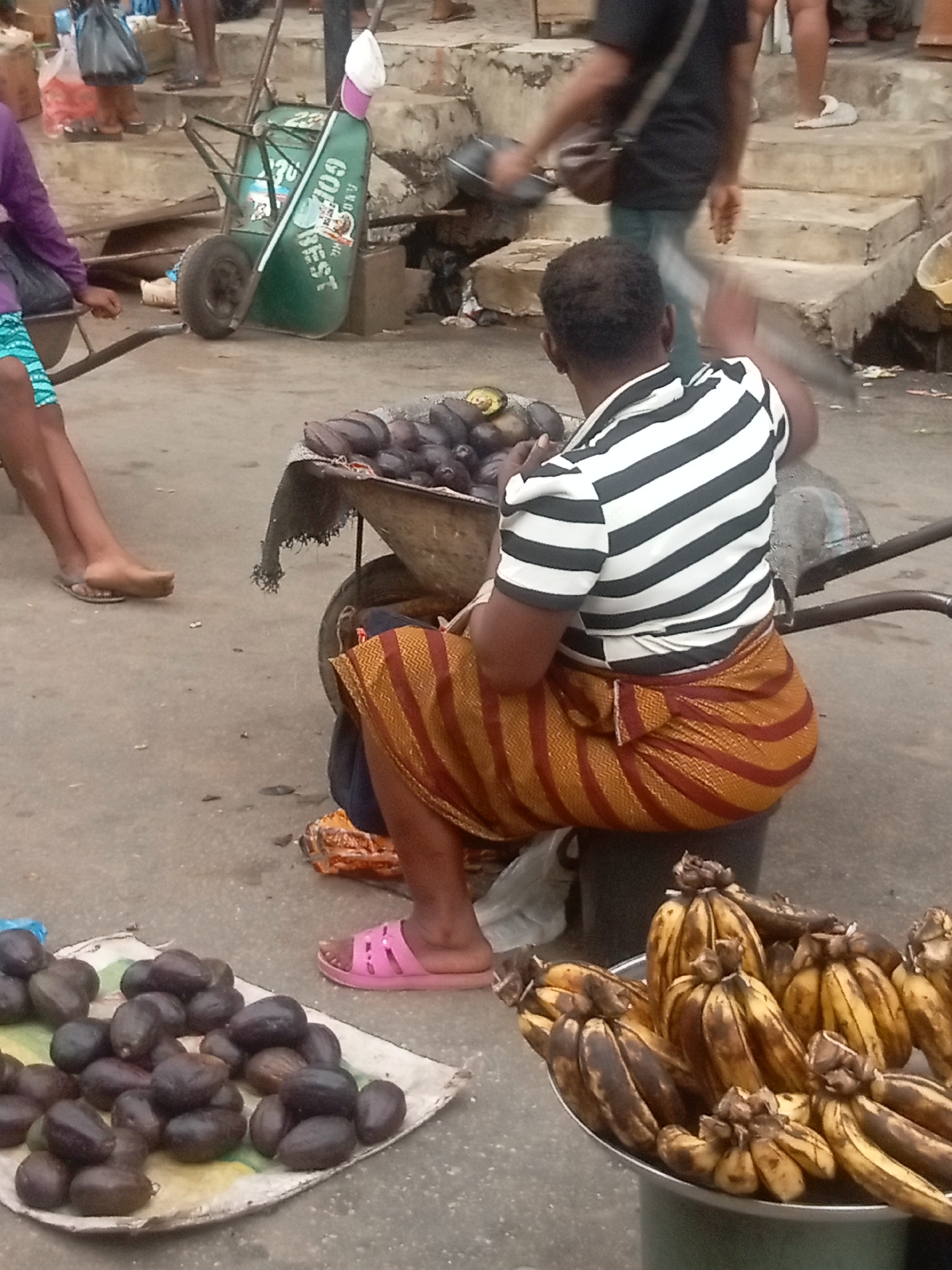 The image captures a scene in Fegge, Nigeria, featuring a woman seated on a gray container, possibly a bucket, at what appears to be a marketplace. She wears a striped black and white shirt and an orange striped wrap-around skirt, and pink sandals. Her back is to the camera. In front of her, a wheelbarrow is filled with dark purple fruits, possibly African pear (Ube). More of these fruits are displayed on a cloth on the ground to the left. A separate tray of bananas sits to the right. Behind her, another wheelbarrow and glimpses of other people and the marketplace setting are visible.