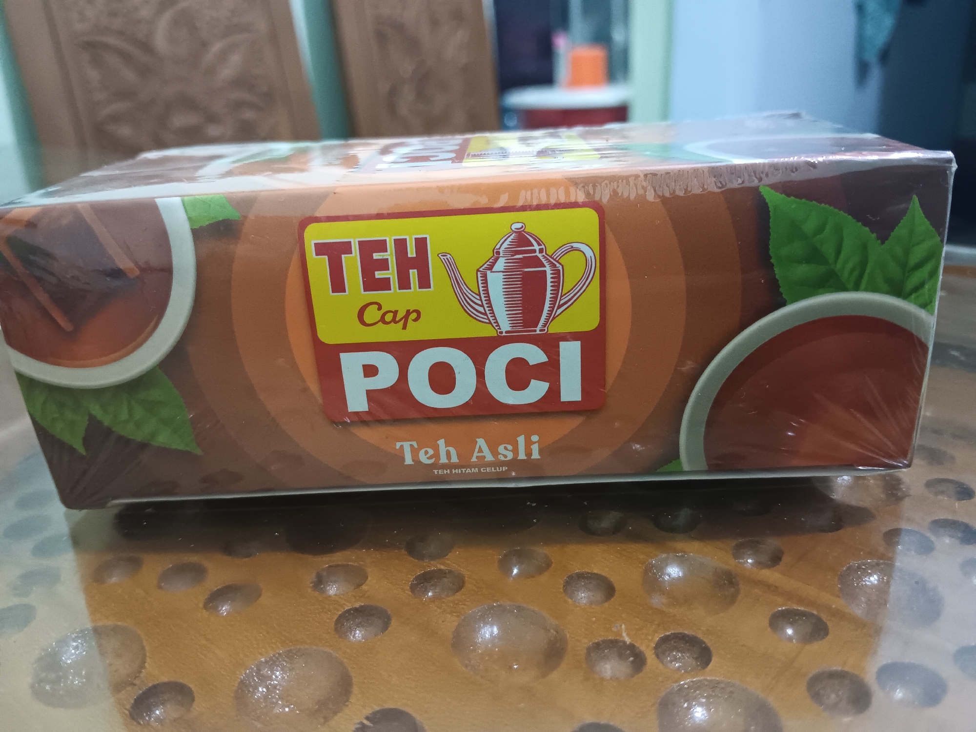 The image shows a close-up of a box of Teh Poci tea, a popular brand in Indonesia. The box is wrapped in plastic and is placed on a wooden surface that appears to be a table. The table has a patterned texture with circular indentations, some of which are reflecting light. The background is blurred, but it seems to be an indoor setting with some furniture, possibly chairs, visible. The lighting suggests it is daytime. The box itself features a prominent yellow label with red text that reads "TEH Cap POCI". There is also an illustration of a teapot and some green tea leaves. Below the main label, smaller text reads "Teh Asli" and "TEH HITAM CELUP". The overall presentation is that of a product shot, possibly for commercial purposes.