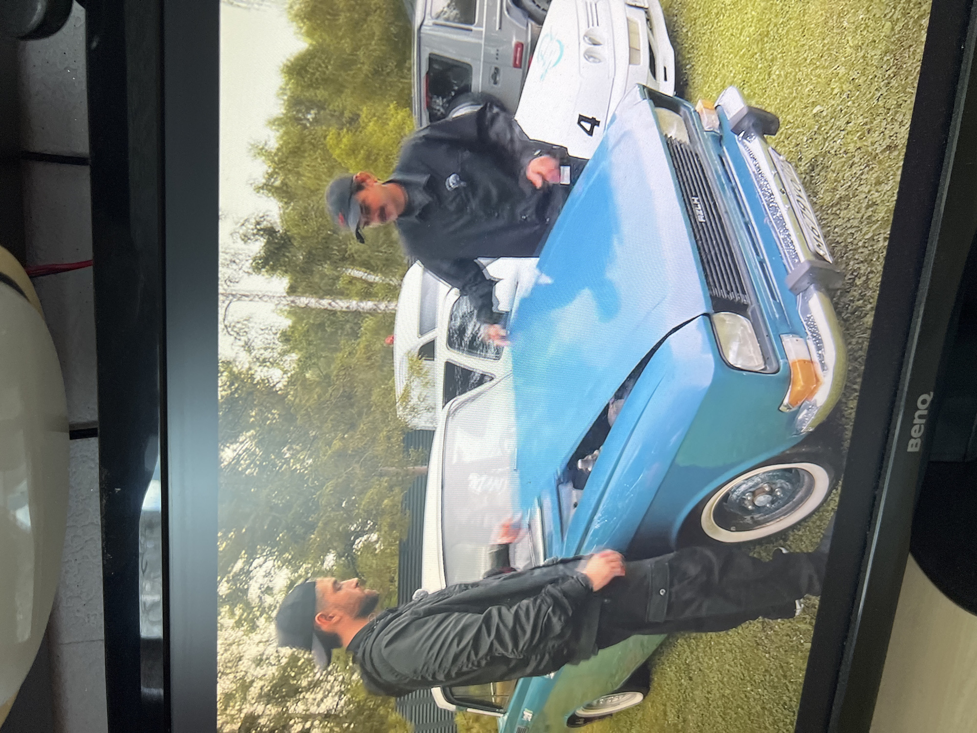 Two men are gathered around a light blue classic car with its hood open. The man on the left, wearing a black baseball cap, black bomber jacket, and black cargo pants, is looking at the car's engine. The man on the right, wearing a dark baseball cap, dark jacket, and dark pants, is reaching towards the open hood. The car has a white roof and white-wall tires. In the background, other vehicles are visible, including a white van and a silver SUV. The scene appears to be set outdoors on a grassy area with trees in the background. The BenQ logo is visible on the bottom of the frame, indicating the image is displayed on a monitor. The license plate on the blue car is obscured, but appears to read "NG 7910". There is no indication of the specific location in the city of Kanash, Russia.