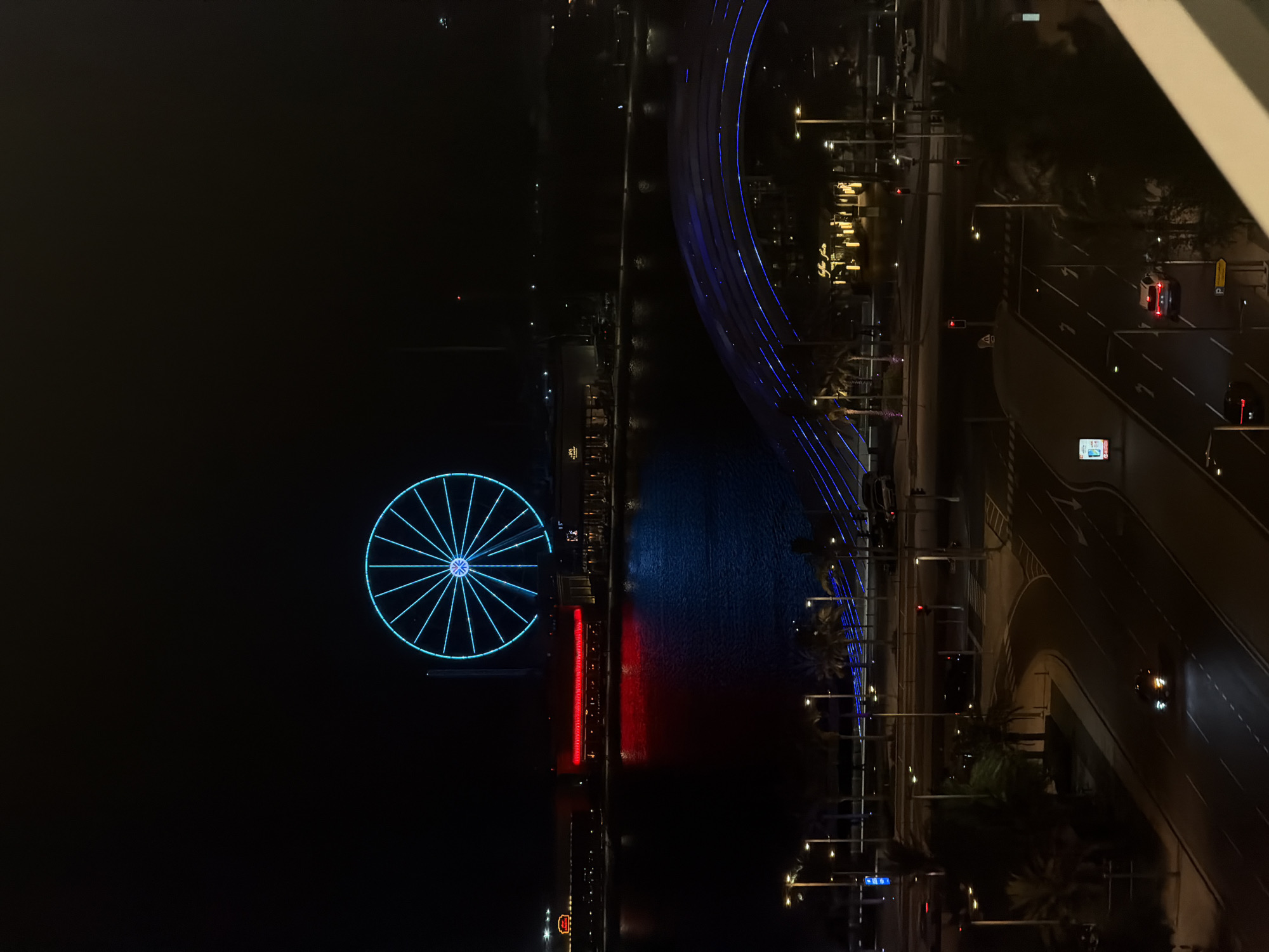 In an aerial nighttime shot of a waterfront cityscape, a large, illuminated Ferris wheel with bright blue spokes dominates the upper left quadrant against a dark sky. Below it, a body of water reflects the red lights of a building on its far shore, creating a distinct red and blue color split on the water's surface. In the foreground, a multi-lane road winds through the scene, illuminated by streetlights and the headlights of passing cars. A modern pedestrian bridge with blue lighting arches across the lower middle of the frame. Palm trees are scattered throughout the foreground and mid-ground, adding to the tropical ambiance. The overall scene depicts a vibrant urban environment at night, with a focus on the illuminated structures and the reflective water.