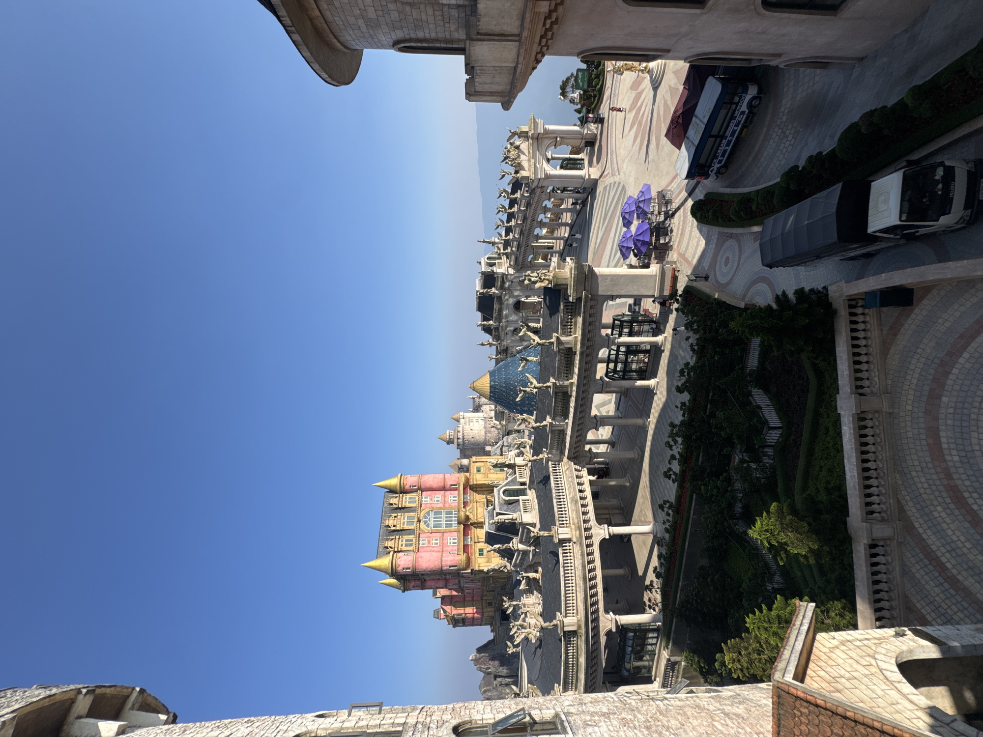 The image provides an elevated, wide view of the French Village at Ba Na Hills, a resort complex situated in the Truong Son Mountains, approximately 25 kilometers southwest of Da Nang, Vietnam. The scene is illuminated by bright daylight under a clear, cloudless blue sky, with distant hazy mountains visible on the horizon.
Architecturally, the complex features numerous stone-clad buildings in a European Gothic revival style. A prominent multi-story building in the mid-ground displays pink and gold facades with multiple windows and four golden spires. An elaborate elevated walkway system, supported by classical columns and arches, connects various sections and is adorned with numerous statues of winged figures atop its roofline. A large, blue-tinted geodesic dome is integrated into this walkway.
Paved plazas below feature intricate tile patterns. Outdoor seating areas with purple umbrellas, tables, and chairs are present. A winding asphalt road traverses the lower right portion of the image. A white Isuzu truck with a dark, tarp-covered bed is observed moving on this road. Further along the road, a blue and white bus, labeled "ICE CREAM DRINKS HOTDOG," is parked.
Human presence is minimal; one individual in red attire walks across a far plaza, and another person is seated at an outdoor table. Landscaping includes formal hedges and evergreen trees within planted beds.