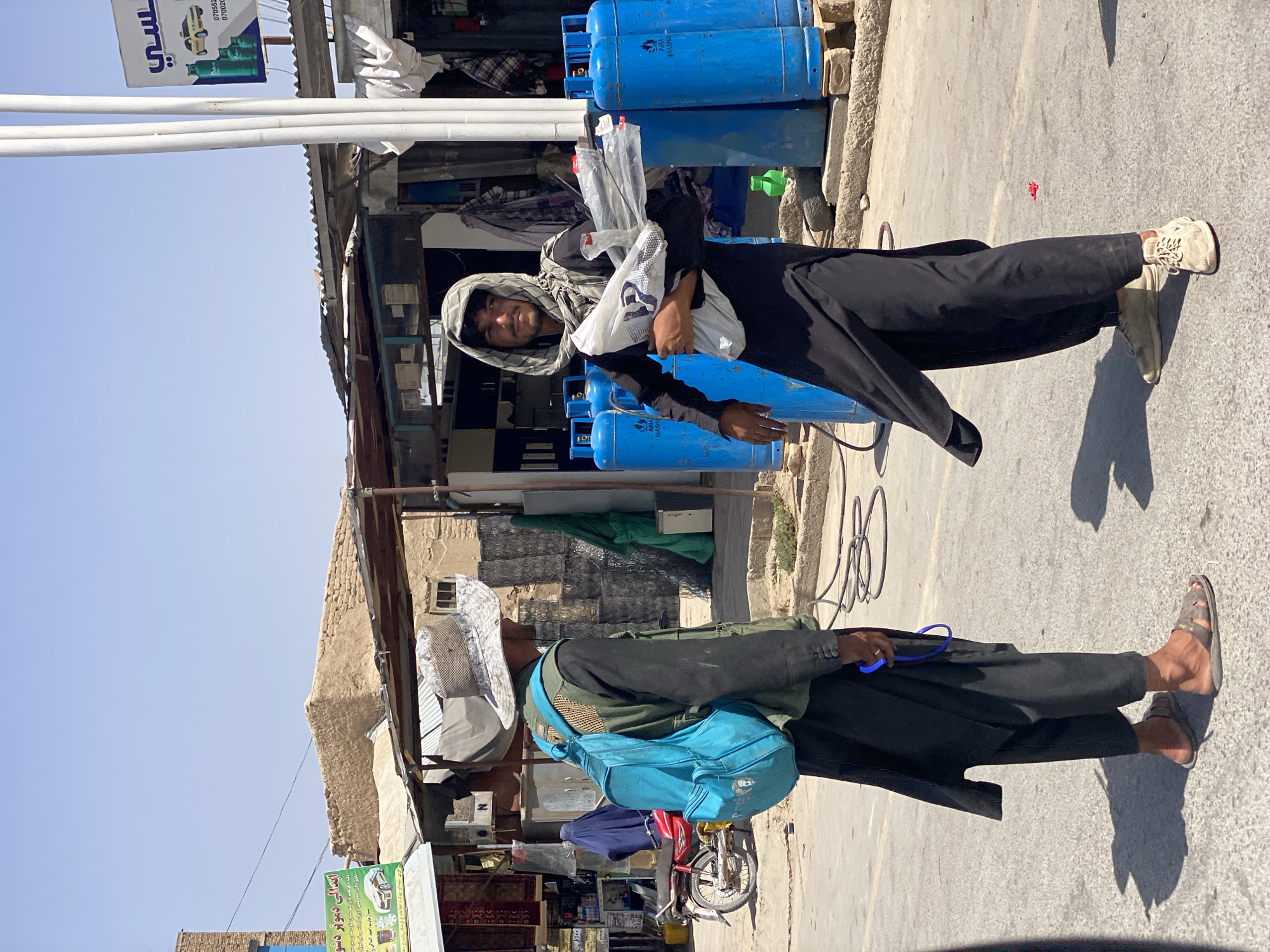 The image depicts two men in what appears to be an outdoor marketplace in the city of Kandahar, Afghanistan, under a bright, clear sky.
The man on the left is wearing a wide-brimmed, light-colored hat, a dark vest over a dark long-sleeved shirt, and dark pants. He has a bright blue backpack on his back and is facing to his right, appearing to be in conversation with the other man. His footwear consists of sandals.
The man on the right is wearing a light-colored checkered headscarf with a dark pattern, a dark long-sleeved tunic, and dark pants. He is holding a white plastic bag filled with what appear to be plastic items, possibly pipes or rods. He is looking towards the viewer. His footwear consists of light-colored sneakers.
In the background, there are several blue gas cylinders stacked together. Further back, there are market stalls with corrugated metal roofs and some merchandise visible, including carpets and other goods. A red motorcycle is parked to the left, and a woman in a blue burqa is visible behind it. The overall environment suggests a bustling, possibly arid, urban setting. The ground is paved with asphalt. A white sign with text and images, including a yellow car and green bottles, is visible on a pole in the right background.