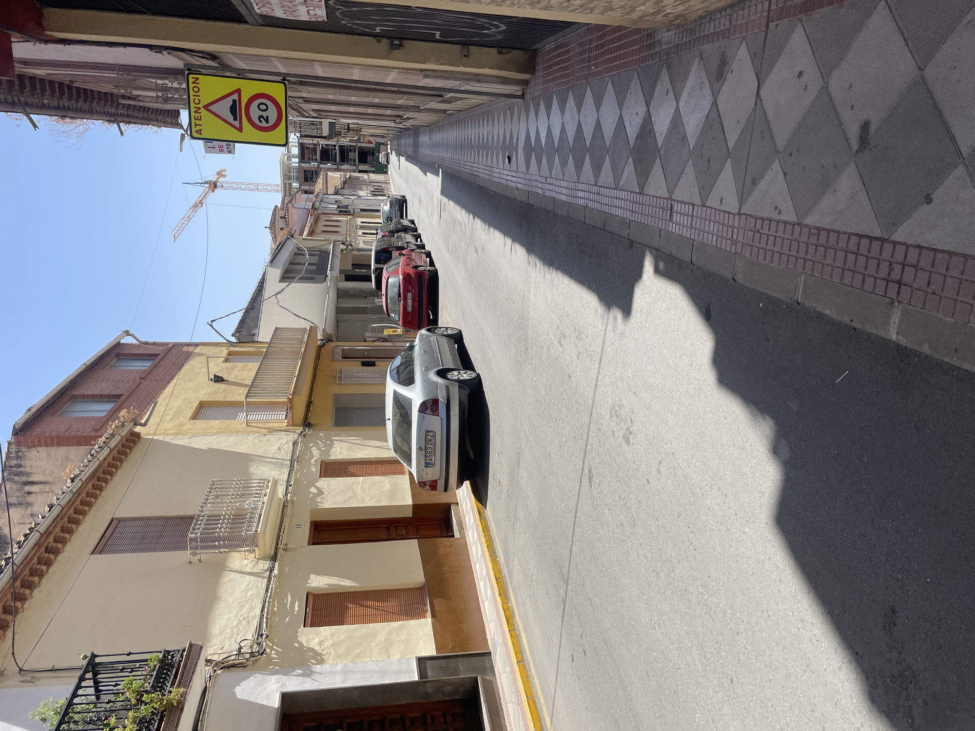 The image depicts a street scene in Atarfe, Spain, on a sunny day. The street is lined with buildings on both sides, with a sidewalk on the right. Several cars are parked along the street and on the sidewalk. A yellow traffic sign with a black border is visible on the right side of the street, indicating a speed limit of 20 and a warning for a speed bump. A construction crane is visible in the background. The buildings appear to be residential and commercial. The sky is clear and blue. The scene is peaceful and everyday.