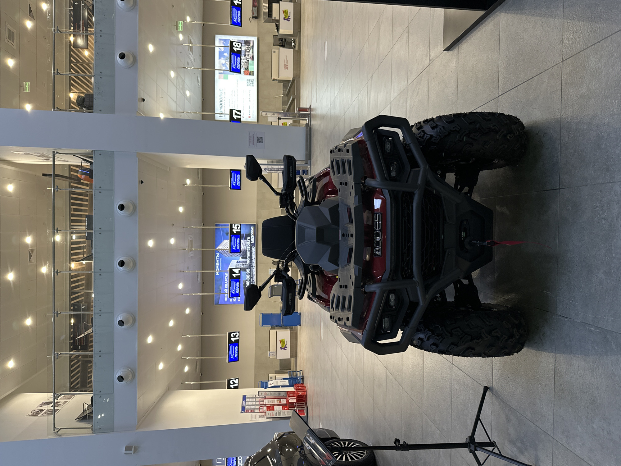 The image depicts the interior of the Perm International Airport (PEE) in Bolshoye Savino, Russia. The scene includes a red and black ATV (all-terrain vehicle) parked in the foreground. Airport check-in kiosks are visible in the background, displaying flight information on screens above them. The kiosks are numbered from 12 to 18. There is a support stand for a camera or similar device. The interior architecture features white columns, security cameras, and lighting fixtures recessed into the ceiling. There are some advertisements.