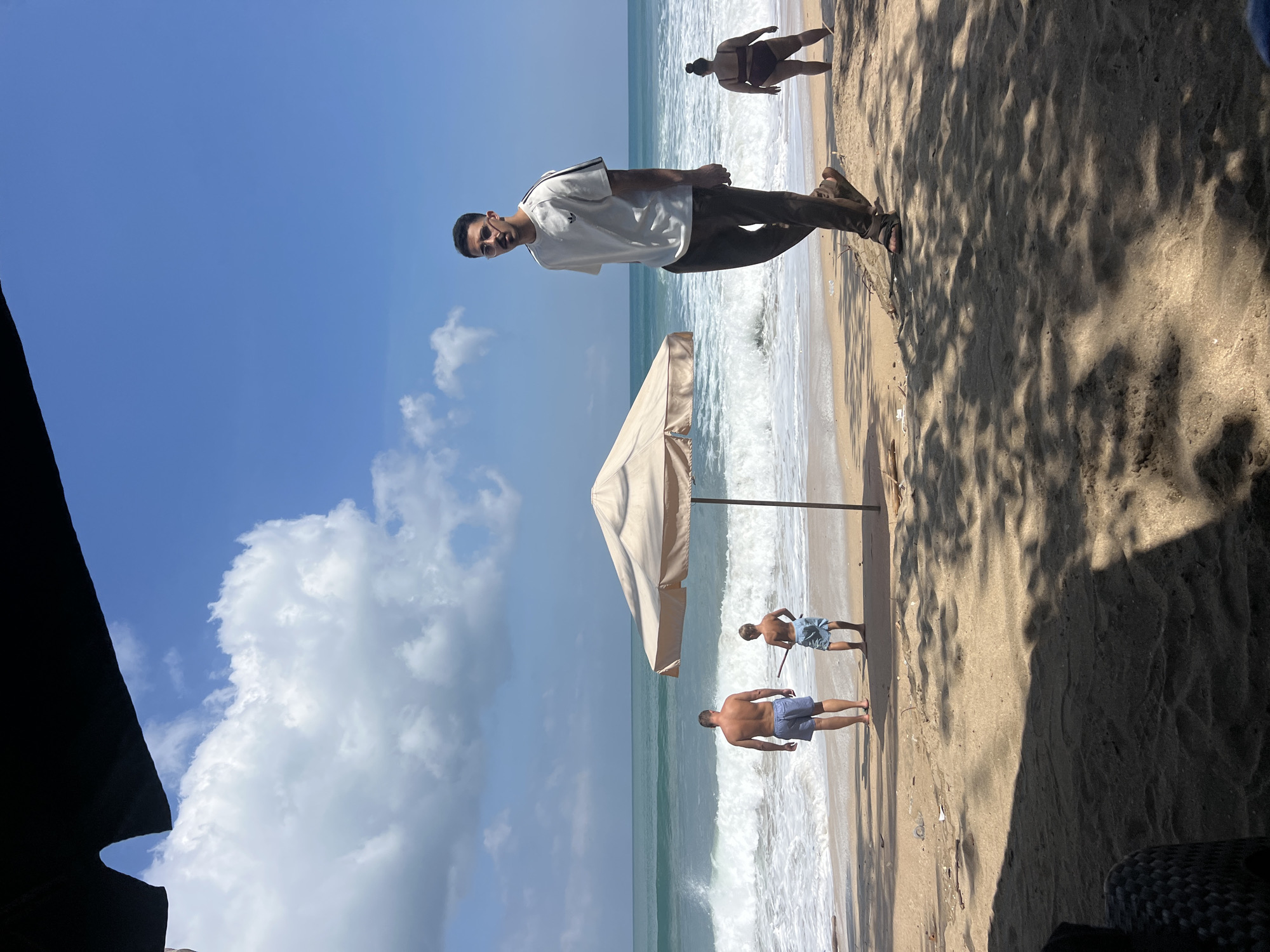 The image captures a sunny day on a sandy beach in Kuta, Indonesia, with the ocean in the background. A man in sunglasses and a white t-shirt with dark stripes on the sleeves is walking towards the camera on the sand. He is wearing dark pants and sandals. In the middle ground, a large, light-colored umbrella stands on the beach. To the left of the umbrella, two individuals are seen facing the ocean. One is a man in blue shorts, and next to him is a younger person in lighter shorts. Further in the background, a woman in a dark swimsuit is walking away from the camera towards the right side of the frame. The waves are breaking on the shore, and the sky is blue with scattered white clouds. The sand is speckled with shadows from unseen trees.