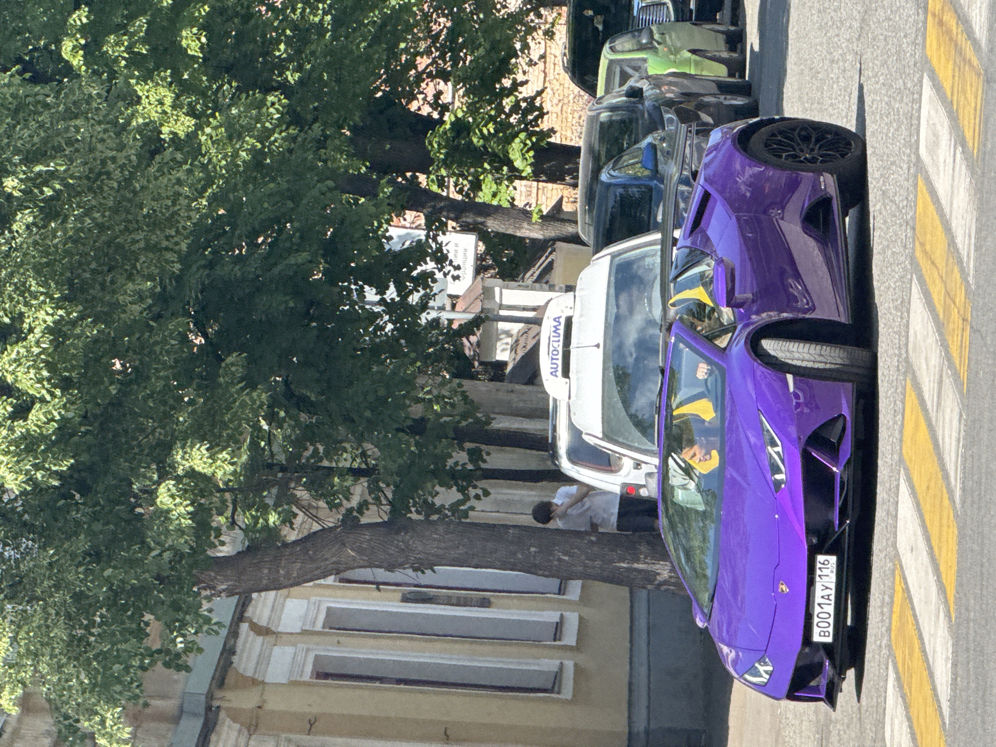 The image shows a purple Lamborghini parked on a crosswalk in Kazan, Russia. The license plate reads "B001AY 116 RUS." Other vehicles, including a white van with "AUTOCLIMA" on the side, are parked along the street. A person in a white shirt stands near the van. Trees line the street, and a building with light yellow walls and white-framed windows is visible in the background.