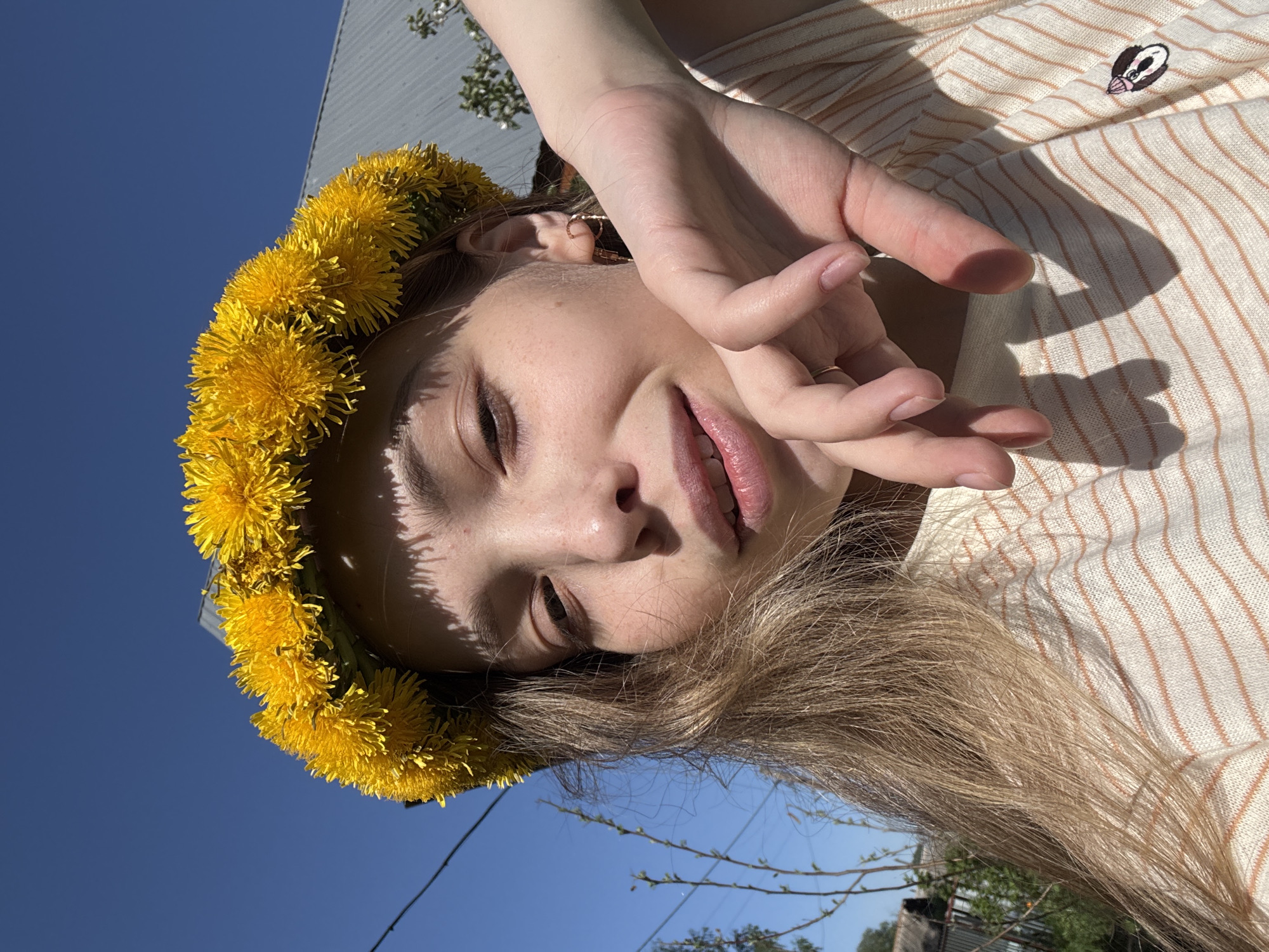 A young person with fair skin and light brown hair is lying down, wearing a crown of yellow dandelions. The person is wearing a striped shirt and has their hand raised near their face. The sky is blue and visible in the background, along with trees and buildings. The person is facing the sky and it is daytime. Кемуль, Russia.