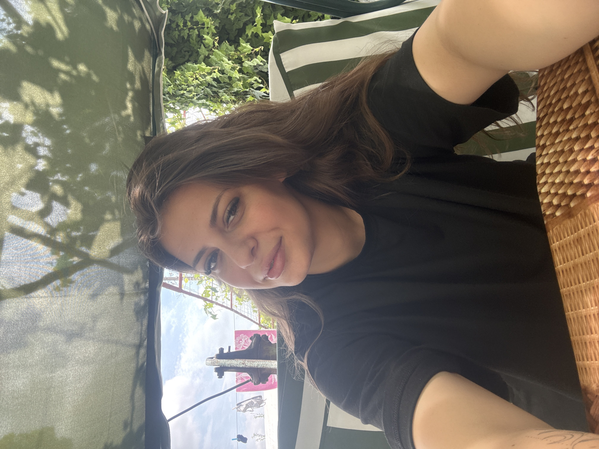 A young woman with long, brown hair, wearing a black t-shirt, is taking a selfie while sitting on a swing set outdoors. She is looking at the camera with a slight smile. The background shows a leafy vine, a striped cushion on the swing, and a glimpse of the sky with clouds. In the distance, there is a metal structure, pink fabric, and laundry hanging on a line. The scene appears to be in a residential area, possibly a backyard, in the village of Govedare, Bulgaria.