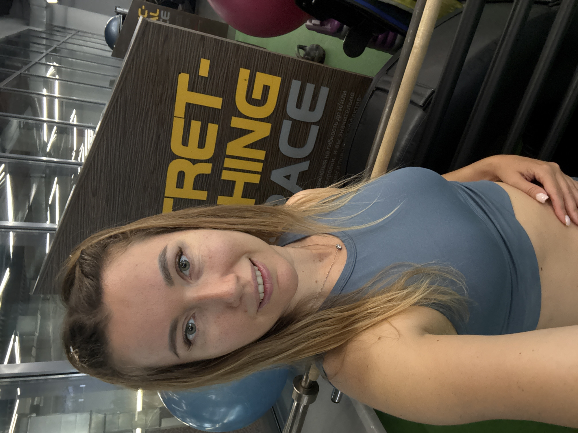 The image is a selfie of a young woman indoors, possibly in a gym or fitness center. She has fair skin, green eyes, and blonde hair. She is wearing a blue sports bra. In the background, there's a sign with the words "STRETCHING SPACE" written on it in yellow and gray letters. The sign appears to be made of wood. Behind the sign, there are gym equipment and some weights. The floor is green, and a window is visible with a view of another building. The lighting appears to be from artificial sources.