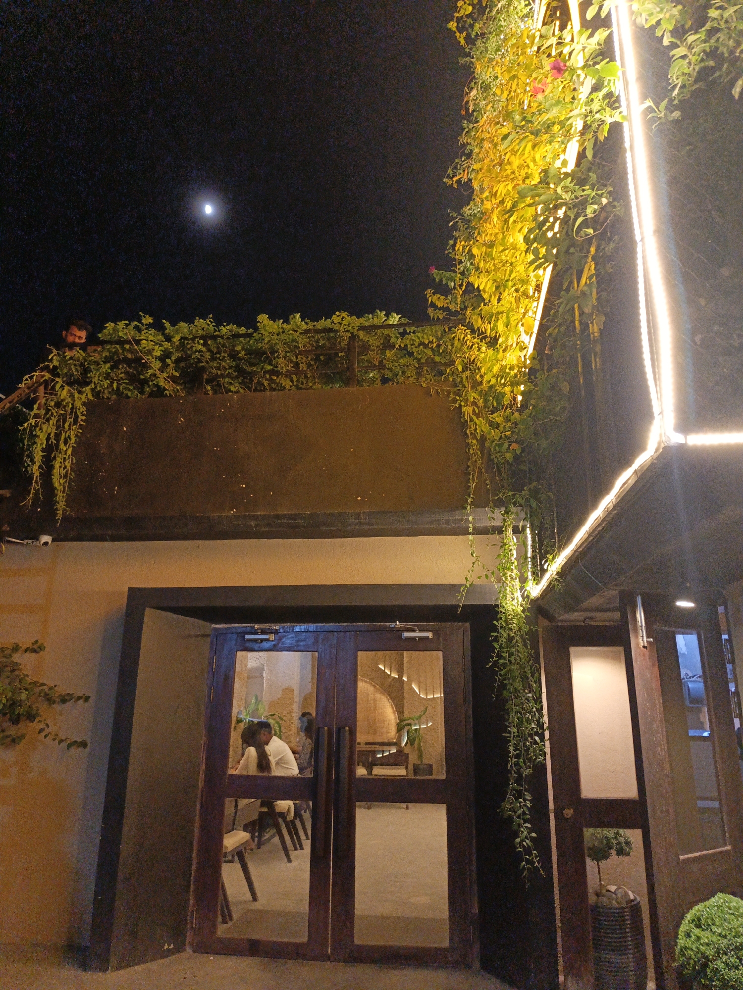 A night exterior shot shows the entrance of a building with a dark facade and wooden doors. Above the entrance, a balcony is adorned with lush green plants and hanging vines, illuminated by warm, bright lights. The sky above is dark, with a visible crescent moon. A man is partially visible peeking from behind the greenery on the balcony. Through the glass doors, patrons are seated at tables inside the establishment. The scene is located in Dhaka, Bangladesh.