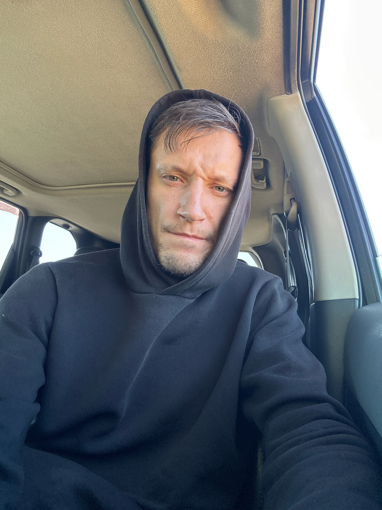 In the city of Makiv, Ukraine, a man with fair skin, short brown hair, and a light beard is photographed from the chest up, inside what appears to be a car. He is wearing a black hooded sweatshirt, with the hood pulled up over his head. His expression is serious, with his brow furrowed. The background shows the interior of the car, including the headliner and a portion of the car's side. The lighting suggests it is daytime.
