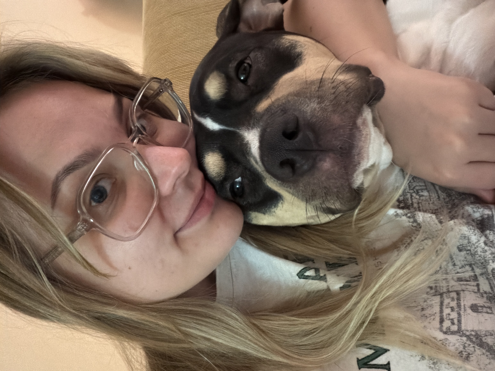 A fair-skinned woman with blonde hair and blue eyes, wearing glasses, is lying down next to a black-and-tan dog. The dog is resting its head on the woman's shoulder. The woman is wearing a t-shirt with a visible portion of the word "N" printed on it. They are resting on a beige couch. The location in the city of Tel-Aviv, Israel is unknown.
