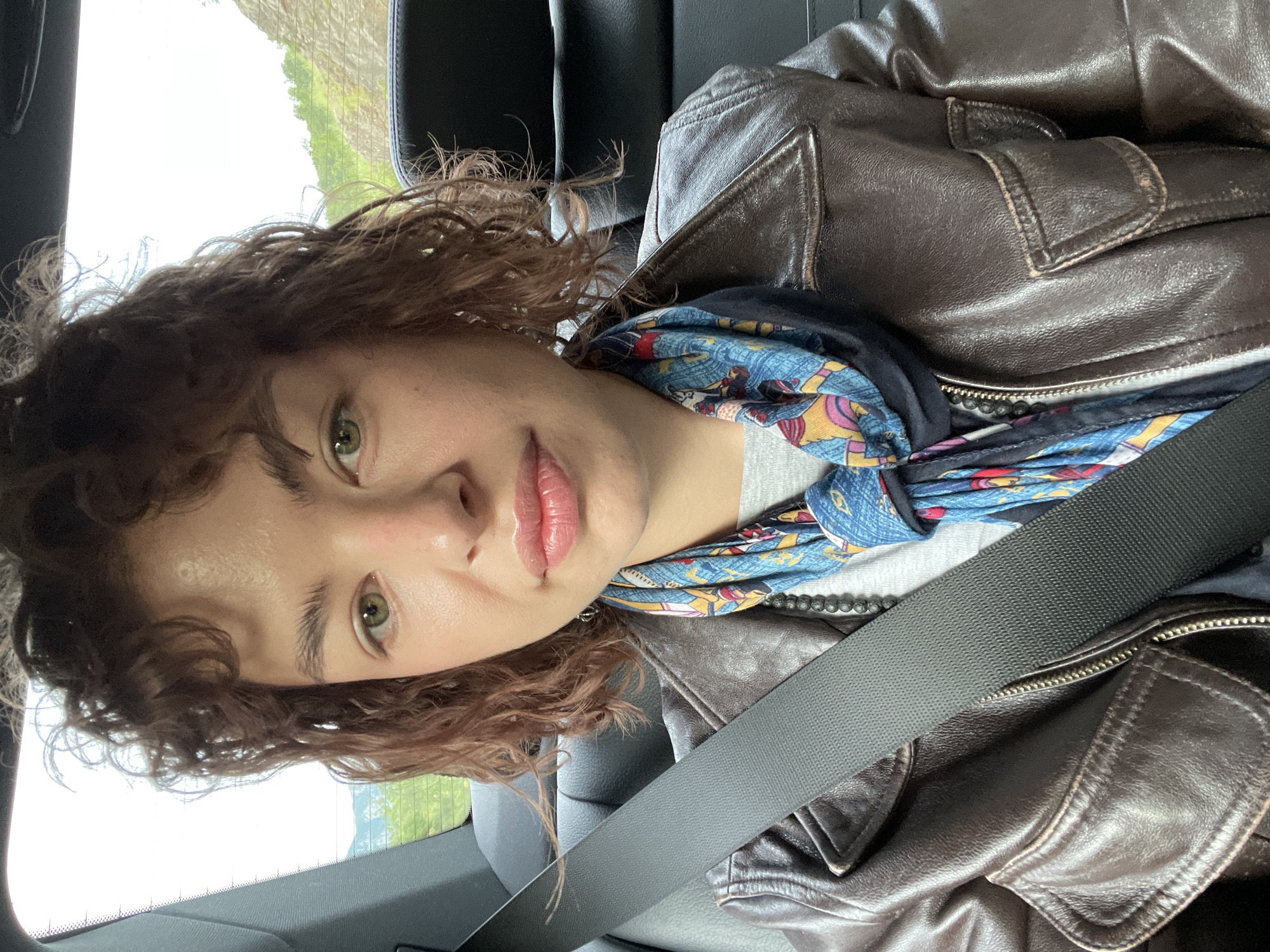A person with curly brown hair and green eyes is in a car. They are wearing a brown leather jacket and a colorful patterned scarf, secured by a seatbelt. Outside the car window, vegetation is visible. The location is Sotonići, Montenegro.