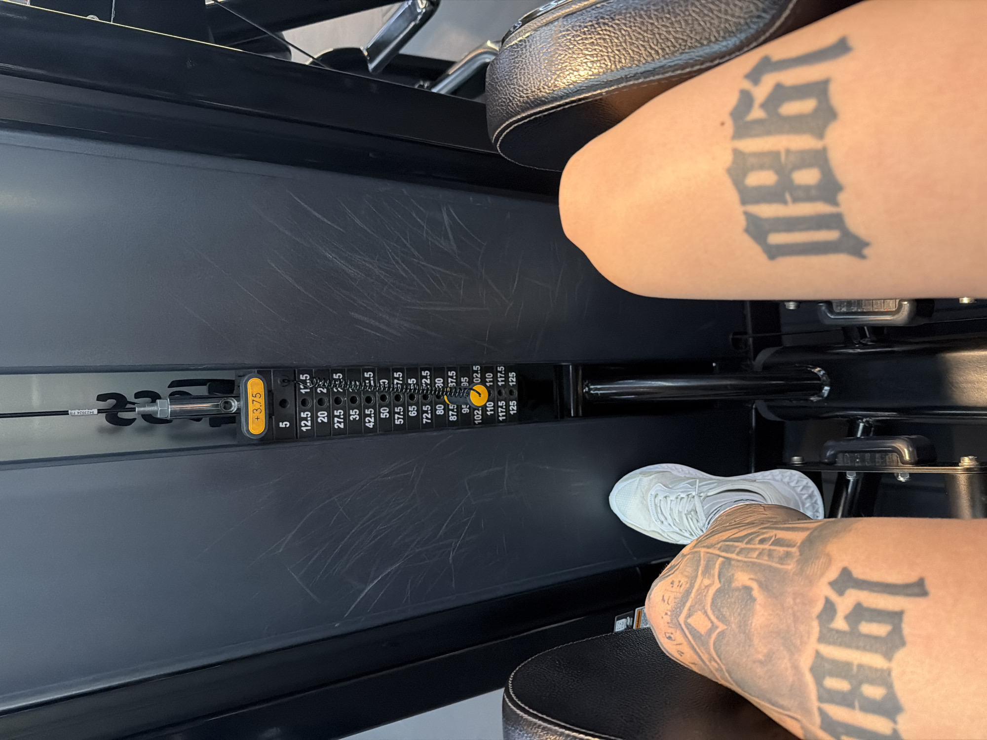 The image captures a close-up, high-angle view of a person's lower body engaged with a weight machine in a gym setting in Plovdiv, Bulgaria. The subject's legs are visible, with the thighs resting on padded supports and the right foot, clad in a light-colored sneaker, positioned on a footrest or lever of the equipment. Both upper legs feature prominent tattoos displaying the stylized numbers "1980". The machine, likely a leg extension or leg press, is dark-colored with visible scuff marks on its surface.
The weight stack of the machine is in focus, showing various weight increments. A yellow pin is inserted into the slot corresponding to "110" units, and an additional smaller yellow pin, labeled "+3.75", is also attached, indicating a total weight of 113.75 units. The weight plates are labeled with numbers such as 5, 12.5, 17.5, 20, 25, 27.5, 35, 42.5, 50, 57.5, 65, 72.5, 80, 87.5, 95, 102.5, 110, 117.5, and 125. The scene depicts an individual actively engaged in a strength training exercise, with the focus on the equipment and the tattooed legs. No specific time of day or emotional state is discernible.