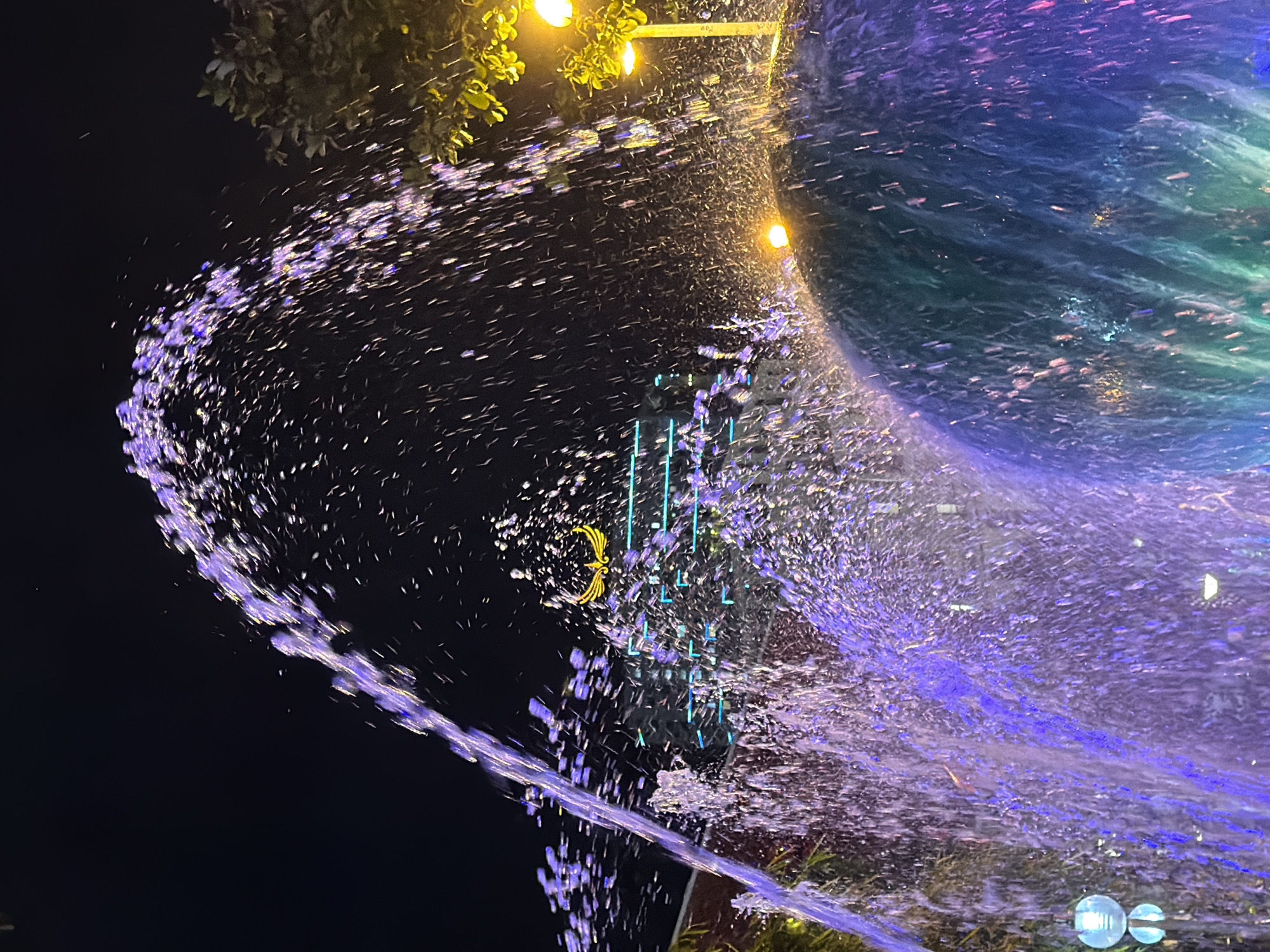 This media file captures a dynamic water fountain display at night, likely in Nha Trang Ward, Vietnam. The primary subjects are the water jets and spray, illuminated by colorful lights, creating a mesmerizing visual effect. The scene is dominated by the movement of water, forming arcs and cascades of illuminated droplets. In the background, glimpses of trees with yellow lights suggest an outdoor setting, possibly a park or public space. There are also vertical lines of blue lights and a golden emblem, hinting at the decorative elements of the fountain's design. The time of day is clearly night due to the dark sky and the prominence of artificial lighting. The water spray, catching the light, is a vibrant mix of purple and blue, with hints of green in the background, indicating the use of colored illumination in the fountain. The overall impression is one of a lively and visually stimulating nighttime attraction. No people are visible in the frame.