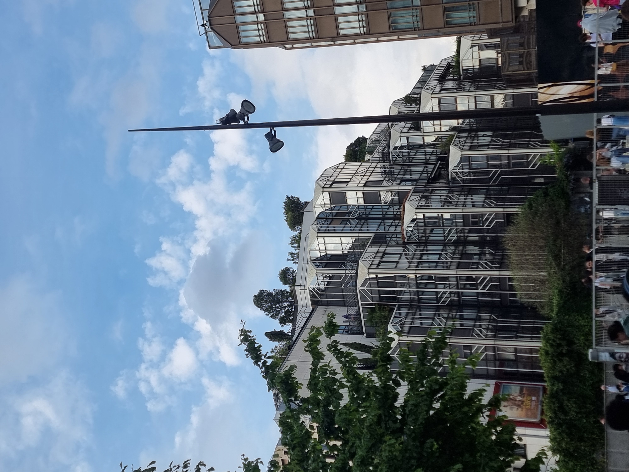 The image shows a view of buildings in Paris, France, under a partly cloudy sky. A tall light pole with two spotlights extends upward. The architectural styles of the buildings vary, including modern glass and metal structures and more traditional buildings with multiple windows. Green trees are visible in the foreground and on the buildings. A crowd of people appears to be gathered near one of the buildings.