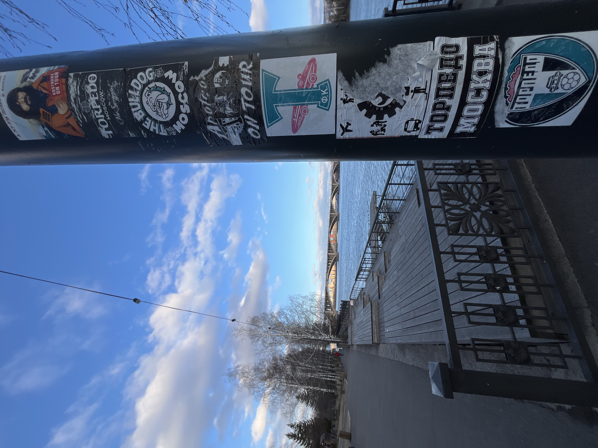 The media file presents an outdoor scene in Krasnoyarsk, Russia, centered on a dark pole covered with various stickers, set against a backdrop of a river and a bright blue sky. The image was taken during the day under sunny conditions, with scattered white clouds visible in the sky. The bare branches of the trees along the riverbank suggest a late autumn, winter, or early spring season.
The main subject is a black pole in the foreground, densely decorated with an array of stickers, many featuring text and graphics related to sports, music, or cultural references. In the mid-ground, a wide river stretches horizontally, with a large arch bridge visible in the distance over the water. A well-maintained wooden embankment or promenade runs along the river's edge, featuring stairs and a decorative black metal railing with intricate patterns. Beyond the promenade, a paved path is visible, lined with deciduous trees (currently bare) and some evergreen trees.
Visible text on the stickers includes:
* "KOSTROMA ON TOUR" accompanying a stylized drawing, along with Russian text "А ХУЛИ ДОМ" and "Ф. Конюхо" (F. Konyukhov).
* "TORPEDO MOSCOW No Surrender"
* "THE BULLDOG MOSCOW" featuring a cartoon bulldog head.
* A prominent black and white sticker reads "ТОРПЕДО МОСКВА" (Torpedo Moscow) in Cyrillic.
* Another emblem contains the word "ТОРПЕДО" (Torpedo).
* A sticker with a large 'T' logo next to the Cyrillic initials "Φ.Κ." (F.K.).
The scene depicts a public recreational area, possibly a riverside park or urban embankment, characterized by local expressions through the graffiti-like stickers.