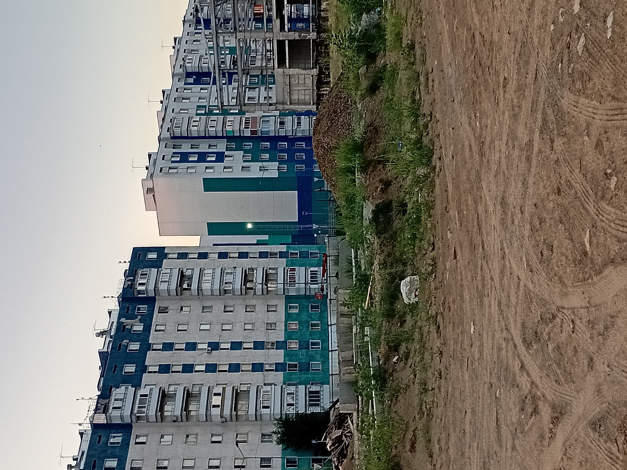 The image depicts a residential construction site in the city of Semey, Kazakhstan. In the background, two large apartment buildings are visible. The building on the left is painted white and dark blue, with multiple balconies. The building on the right is white and green, with a section of exposed concrete frame in front of it, indicating ongoing construction. A large pile of reddish-brown soil or gravel is situated in the middle ground, with construction materials and debris to the left of it. The foreground is an open area of dry, dirt ground with tire tracks visible, suggesting vehicle activity. Green vegetation is sparsely growing on the uneven terrain. The sky is overcast and light-colored, suggesting either dawn or dusk. There are no people visible in the scene.