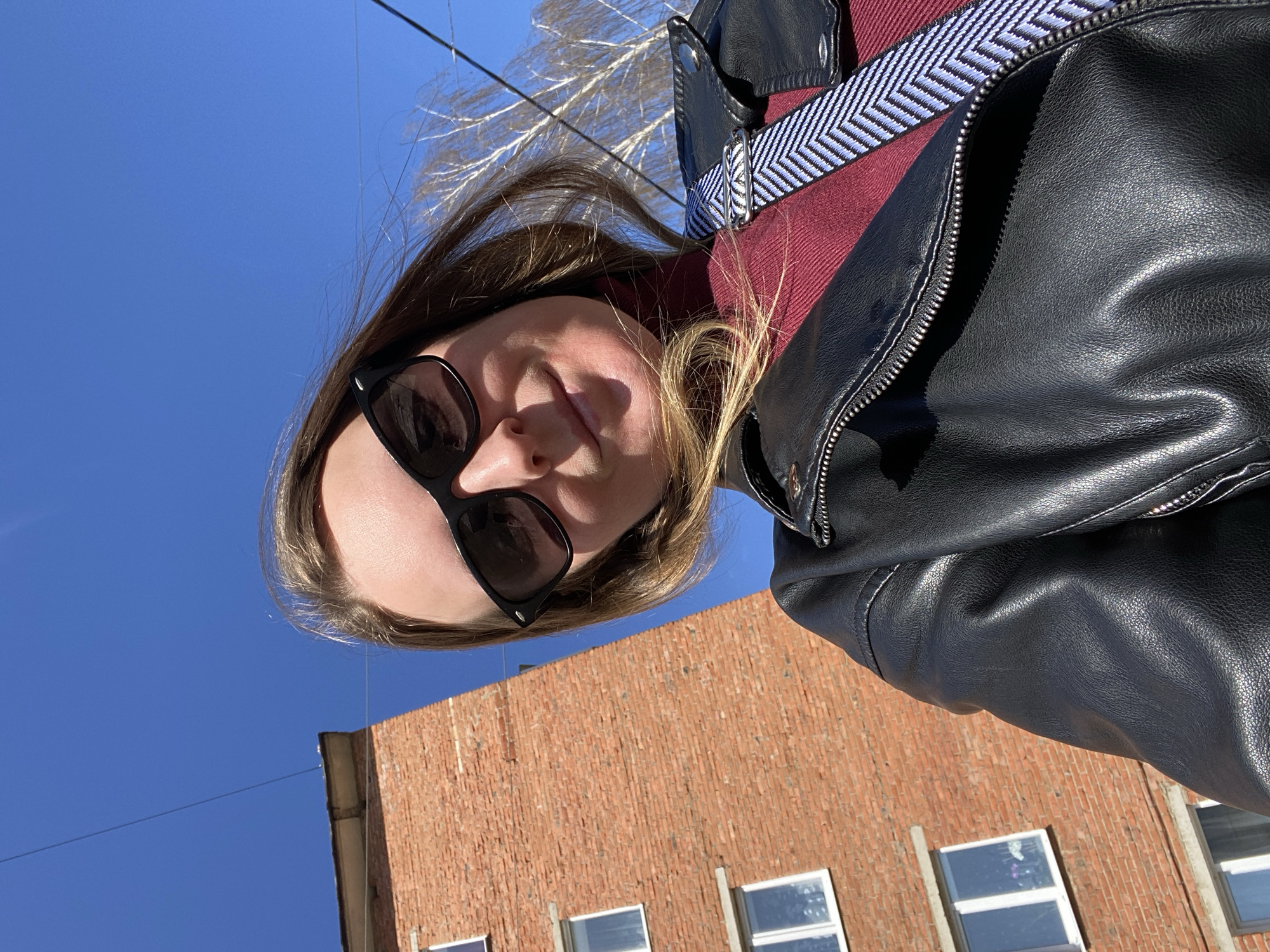 The image shows a person wearing sunglasses, a burgundy sweater, and a black leather jacket. The person is positioned at an angle, looking down towards the camera. In the background, there is a clear blue sky with tree branches visible at the top, and a brick building with windows at the bottom. There is also a wire in the upper left corner. The building suggests an urban environment. No specific information to identify the location as Noviy, Russia is present.