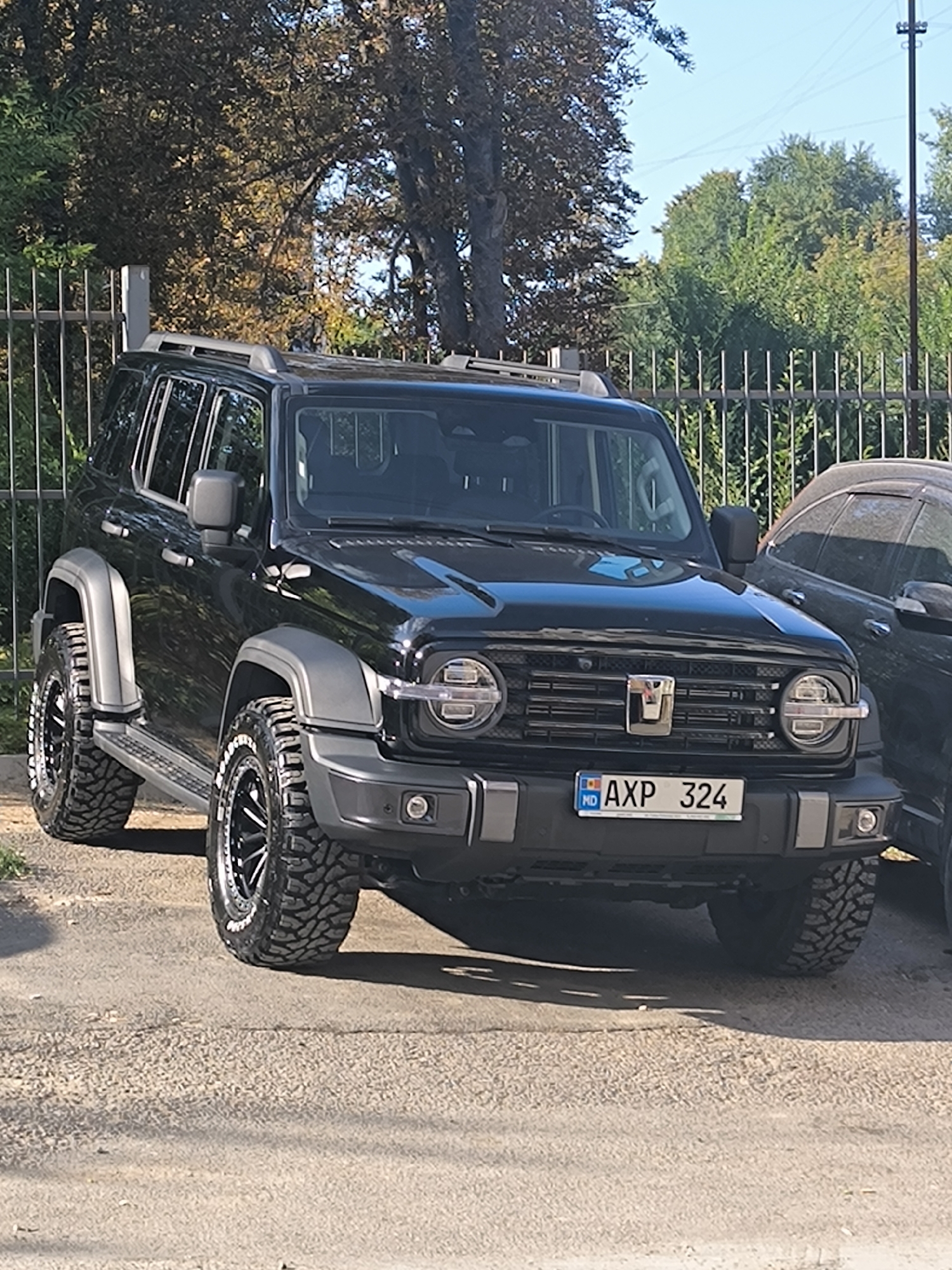 A black SUV, likely a Tank 300, is parked on an asphalt surface in front of a metal fence. The vehicle's license plate reads "AXP 324 MD", indicating it is registered in Moldova. The SUV has off-road tires and appears to be in good condition. Behind the SUV, another dark-colored car is partially visible. The environment suggests an outdoor setting, possibly a parking lot or a residential area, with trees and foliage in the background under a clear sky. The time of day appears to be daytime, with sunlight casting shadows. The image is a frontal view of the SUV, slightly angled. No people are visible in the frame.
