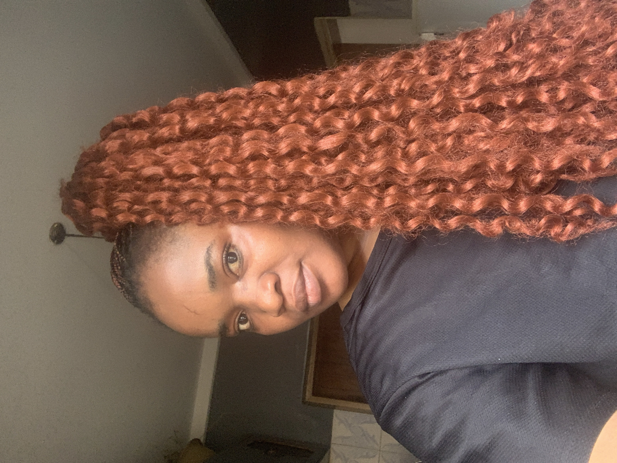A medium shot captures a Black woman indoors, looking directly at the camera with a calm, neutral expression. She has dark eyes, a smooth complexion, and naturally full lips.
Her hair is a prominent feature, styled in very long, voluminous, reddish-brown curls or waves that extend out of the frame. Closer examination reveals visible cornrows near her hairline, indicating the curly hair is likely an extension or a protective braided style.
She is dressed in a dark-colored (presumably black) t-shirt with a subtly textured fabric.
The background depicts an indistinct indoor setting, possibly a room, characterized by plain light-colored walls and some blurry dark elements that might be furniture or architectural details. The lighting is even, suggesting an interior space that is neither overtly bright nor dim.
The woman is posing for the photograph, with no specific activity beyond this. There is no visible text in the image. The location context for the image is Jos, Nigeria.
