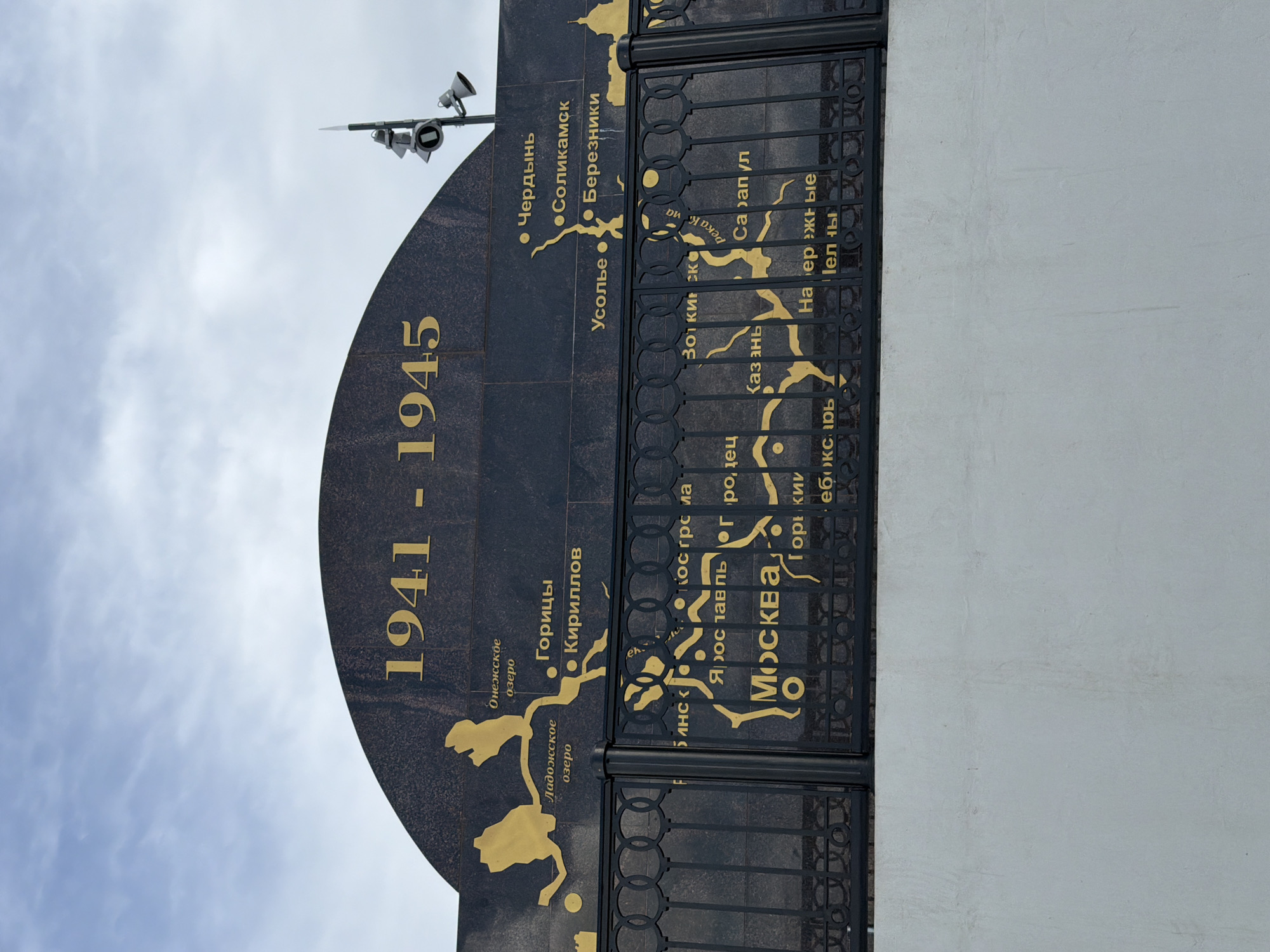 This is a close-up, low-angle, full shot of a memorial structure with gold lettering against a black background, set against a pale blue sky with white clouds. The memorial is a large, dark, curved plaque with gold text and a map-like illustration. The numbers "1941 - 1945" are prominently displayed vertically on the left side of the plaque.
The text, written in Cyrillic, appears to be a list of locations, likely related to historical events. Some legible place names include "Москва" (Moscow), "Казань" (Kazan), "Сарапул" (Sarapul), and "Усолье" (Usolye). There are also references to lakes, such as "Онежское озеро" (Lake Onega) and "Ладожское озеро" (Lake Ladoga). The illustration shows stylized landmasses and bodies of water, connecting these locations.
On the right side of the plaque, a decorative black metal grate with circular patterns obscures part of the lettering and illustration. Above and to the left of the memorial, a metallic structure with multiple speakers or lights is visible against the sky. The lighting suggests it is daytime, possibly morning or afternoon, with diffused light from the overcast sky. The overall impression is one of historical remembrance, possibly commemorating wartime events or routes in Russia. The memorial is positioned outdoors, as indicated by the sky and clouds.
