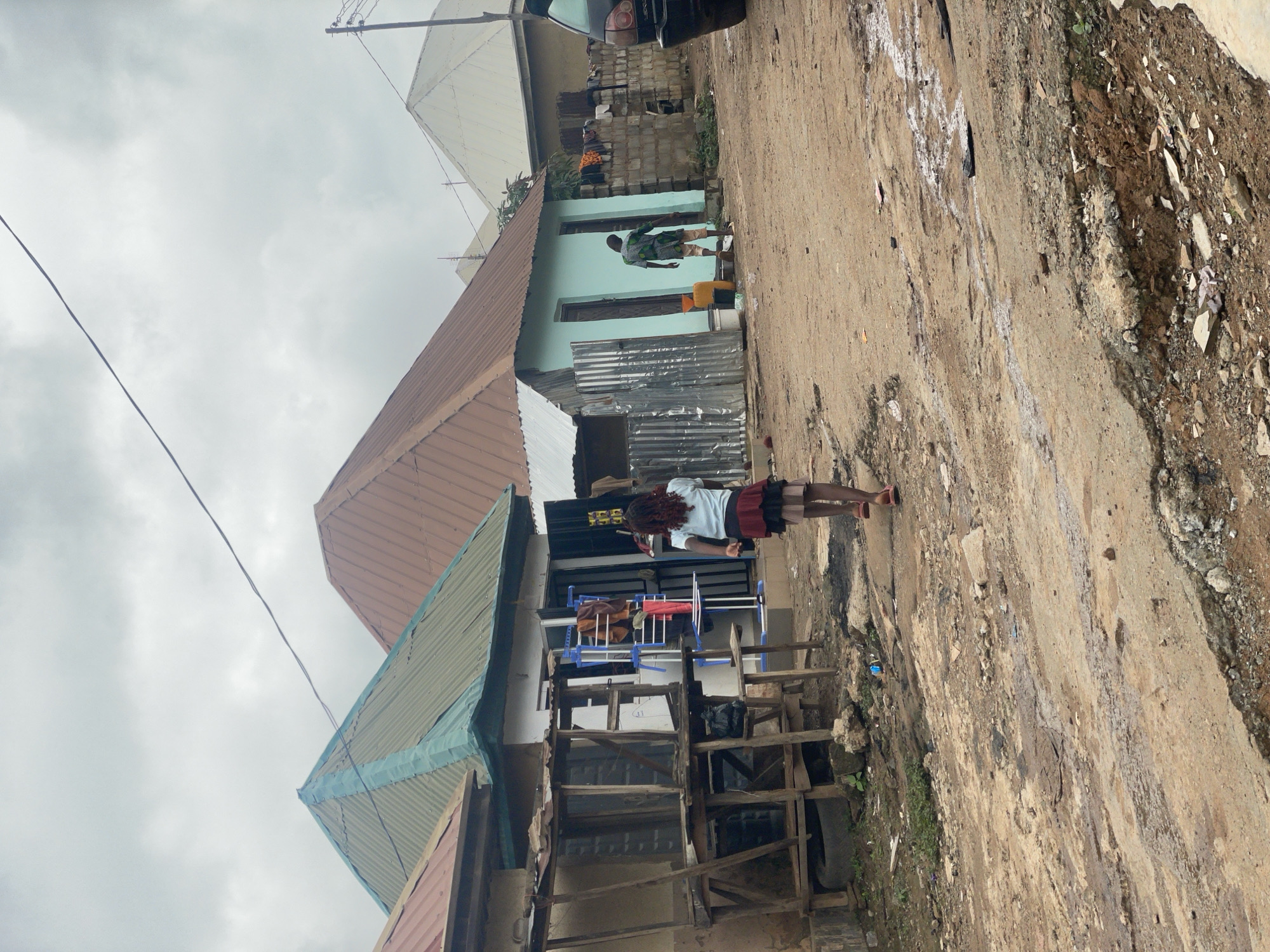 In Kurunduma, Nigeria, under an overcast sky, a dirt road is marred by potholes and puddles, indicating poor road conditions. On the right side of the road, a dark-colored sedan is parked. Further into the scene, a young girl with dark curly hair, wearing a blue t-shirt and a red and black skirt, walks away from the camera on the right. She is walking towards a row of buildings.
To the left of the girl, a modest, light blue building with a brown, corrugated metal roof stands. Its entrance is open, revealing a clothes drying rack with garments. To the left of this building, a more weathered, open-air wooden structure with a green, corrugated metal roof juts out. This structure appears to be a stall or shop, with shelves and some items displayed.
On the far right side of the image, a man dressed in a patterned green and black garment walks away from the blue building. He is heading towards a low wall constructed of concrete blocks, behind which more buildings are visible. The overall impression is a functional, everyday scene in a developing urban area, characterized by rudimentary infrastructure and the daily activities of its residents.