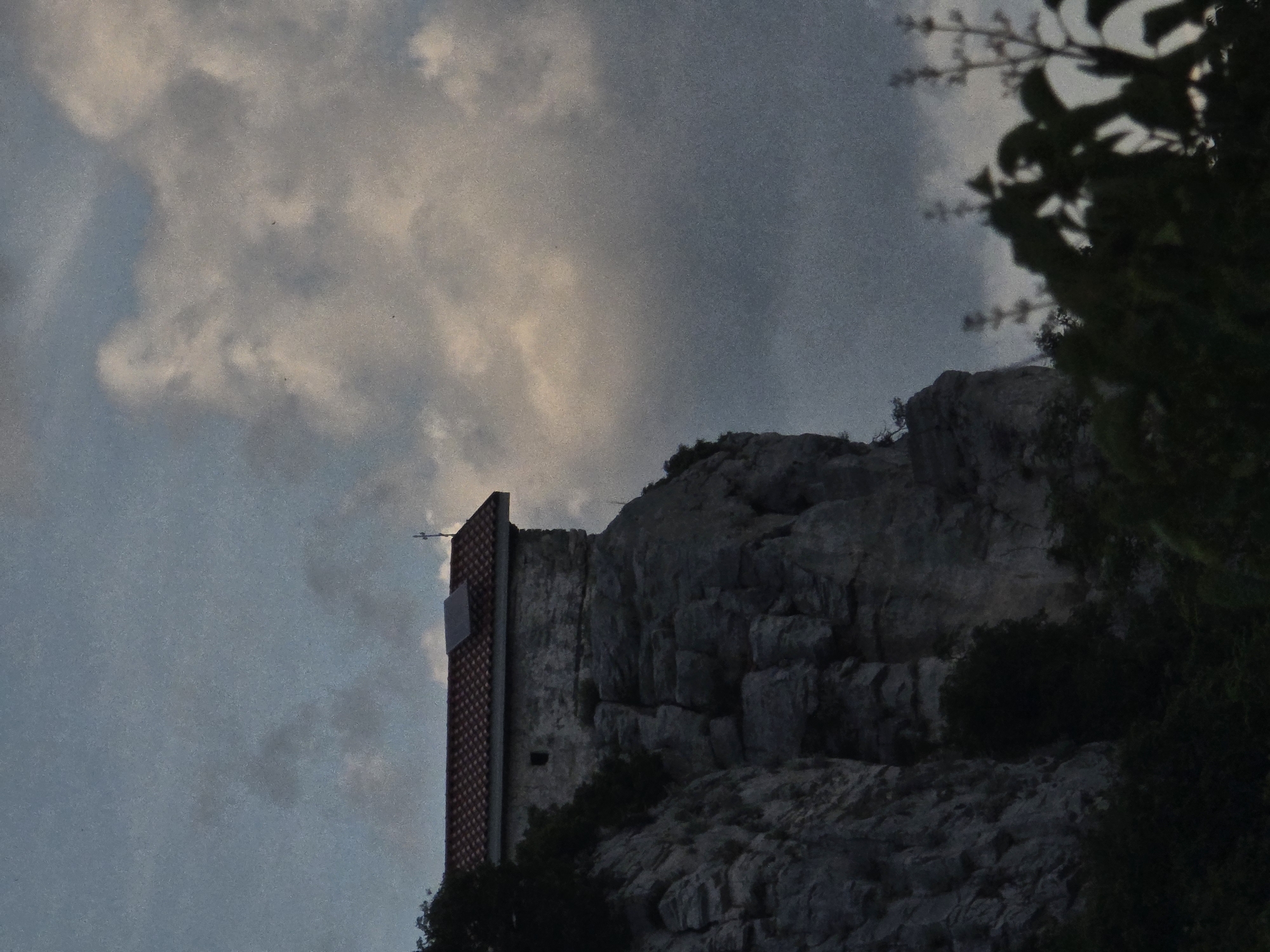 The image shows a stone structure with an adjacent building, possibly in Asenovgrad, Bulgaria, given the prompt's information. The building has a red-tiled side and a white window. The stone structure appears to be a natural rock formation. Vegetation is visible on the right side and at the base of the structure. The sky is cloudy. The scene seems to be captured at dusk or in low-light conditions.