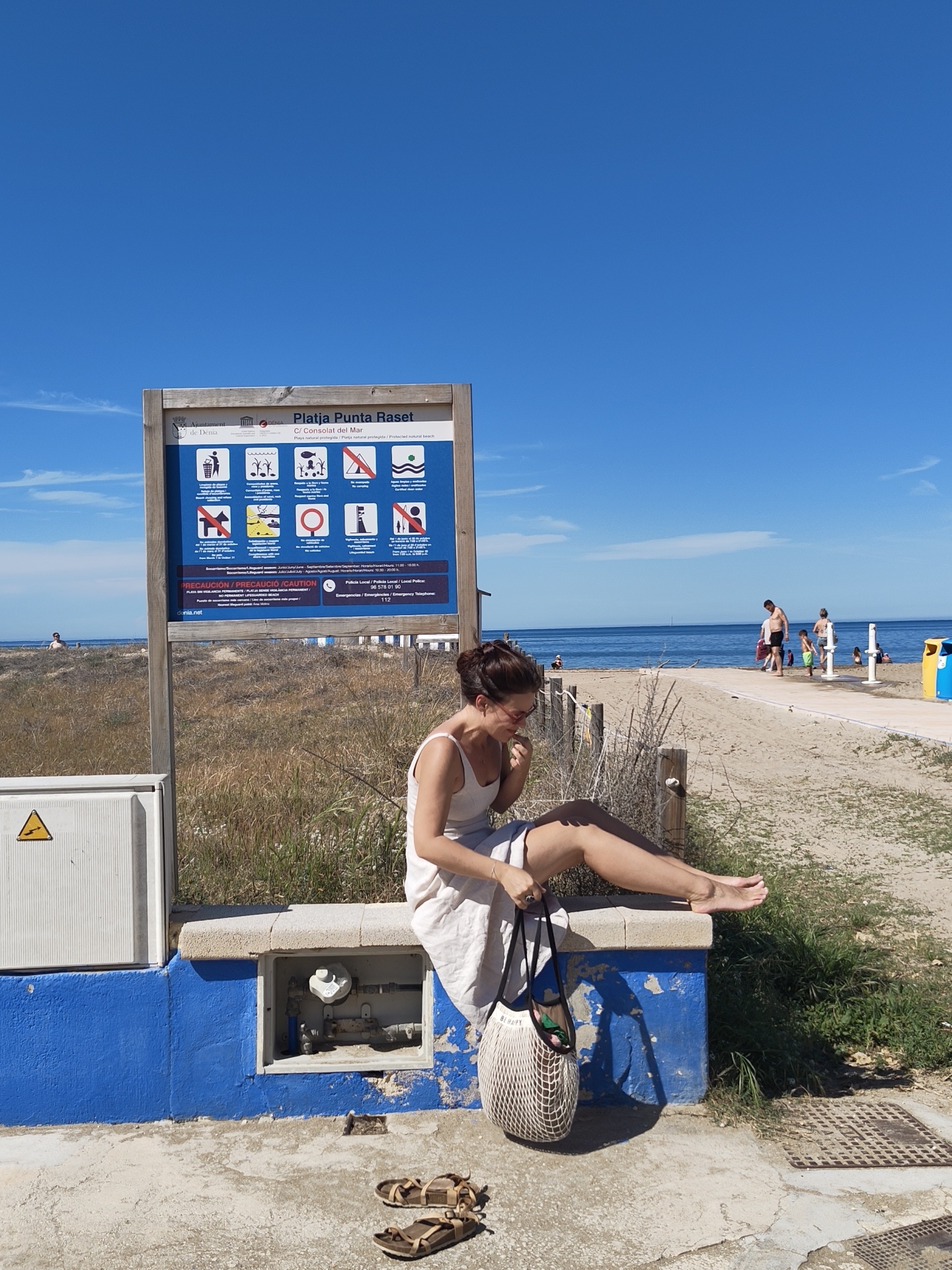 The image shows a woman sitting on a blue-and-white concrete structure near Platja Punta Raset in Dénia, Spain. She is wearing a light-colored dress and holding a mesh bag. Her sandals are on the ground next to her. Behind her is a sign with beach regulations, and in the background, there's a beach with people and the ocean. The sky is clear and blue.